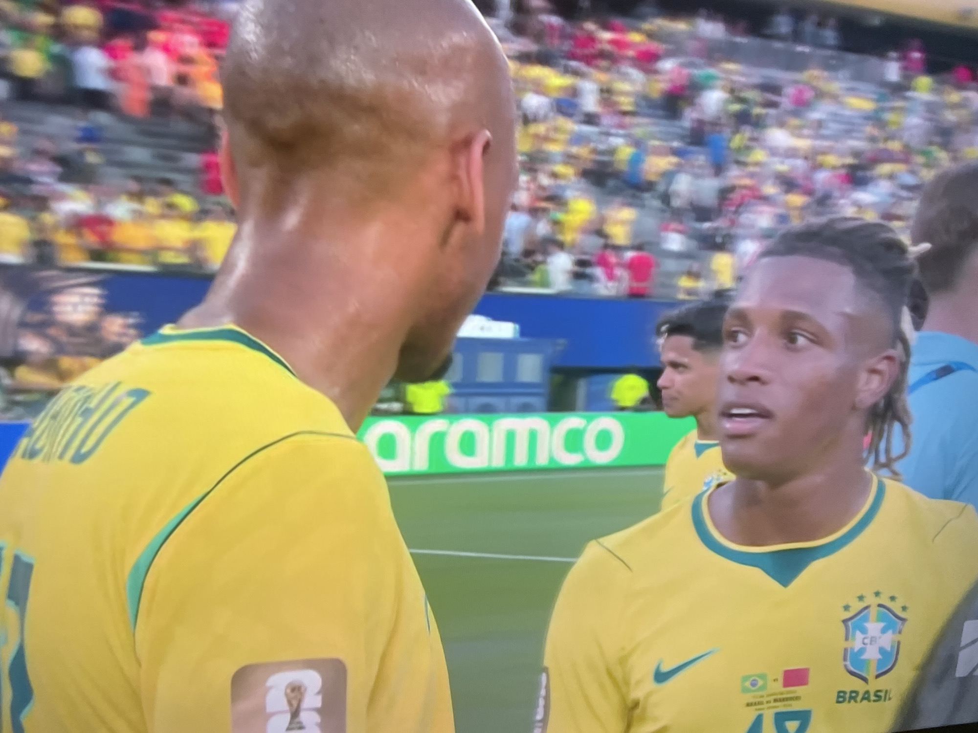 The image shows a close-up of two male soccer players in yellow and green Brazil national team uniforms on a bright, sunny day, likely during a match. The player on the left, partially visible from the neck up and from behind, has a bald head and is sweating. His jersey has teal trim and a patch on the shoulder with a trophy symbol. The player on the right is in profile, looking to his left. He has dark, braided hair and is also sweating. His jersey features the Brazil crest, a Nike logo, and what appears to be a small flag patch with Brazilian and another country's flag. In the background, a blurred crowd of spectators is visible in stadium seating. A bright green advertising board with the word "aramco" in white lettering is also visible. The scene appears to be a soccer field with the players on the grass near the sideline. The image quality is somewhat grainy, suggesting it was taken from a screen or with a less-than-ideal camera. There is no information within the image to suggest the location is in Tbilisi, Georgia.