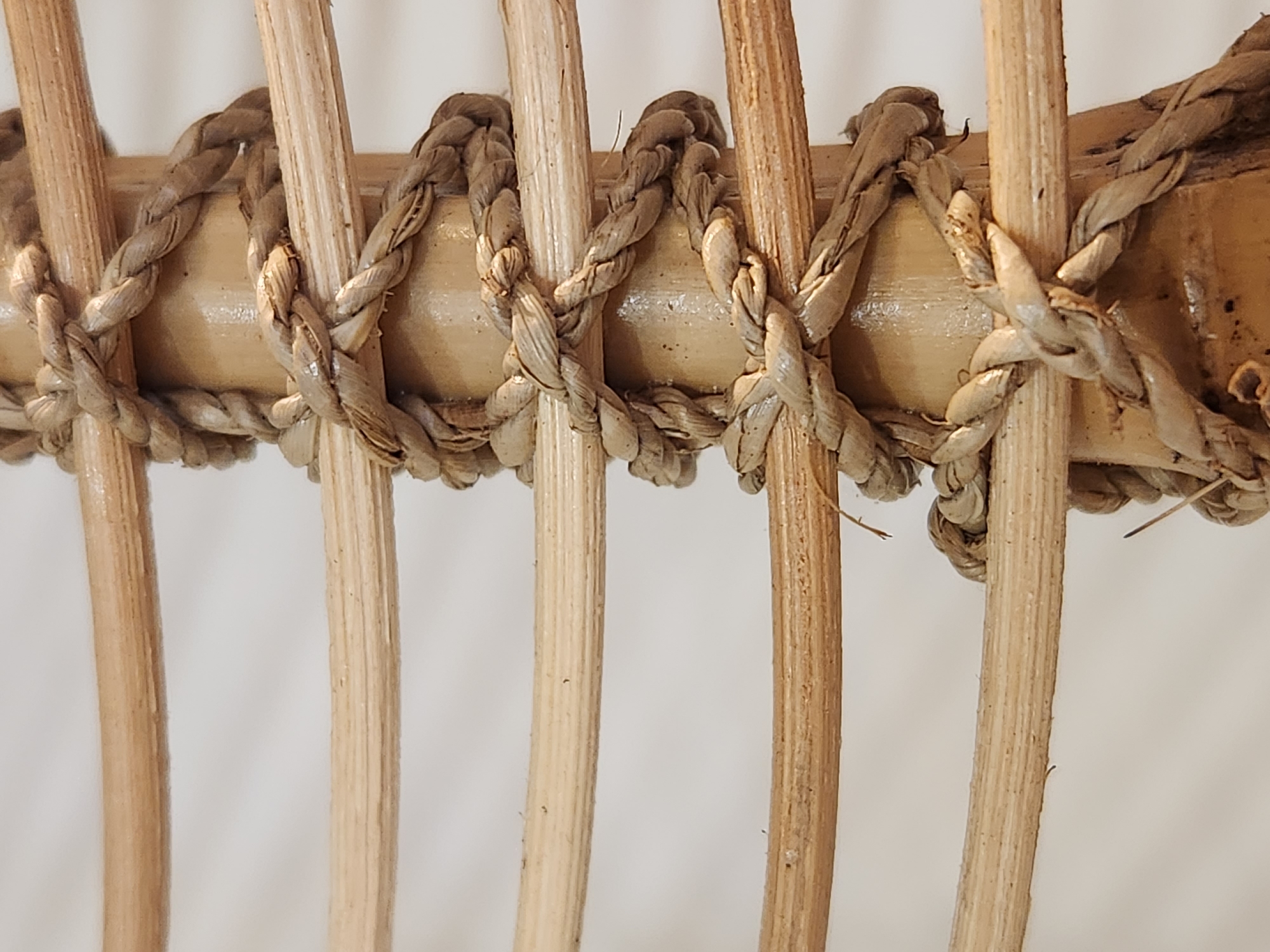 The image is a close-up, vertical shot of natural, light-brown rope intricately wrapped around a vertical, smooth, light-brown bamboo pole. The rope is thick and appears to be made of twisted plant fibers, creating a woven texture. Several horizontal, thin, light-brown bamboo rods are visible, passing behind and in front of the rope and pole, creating a lattice-like structure. The background is a plain, out-of-focus, light-colored surface. There are no people or actions depicted. The scene appears to be a detail of a decorative or structural element, possibly from furniture or a handicraft.
No specific location in Maitland, United States of America, can be determined from the image itself.
