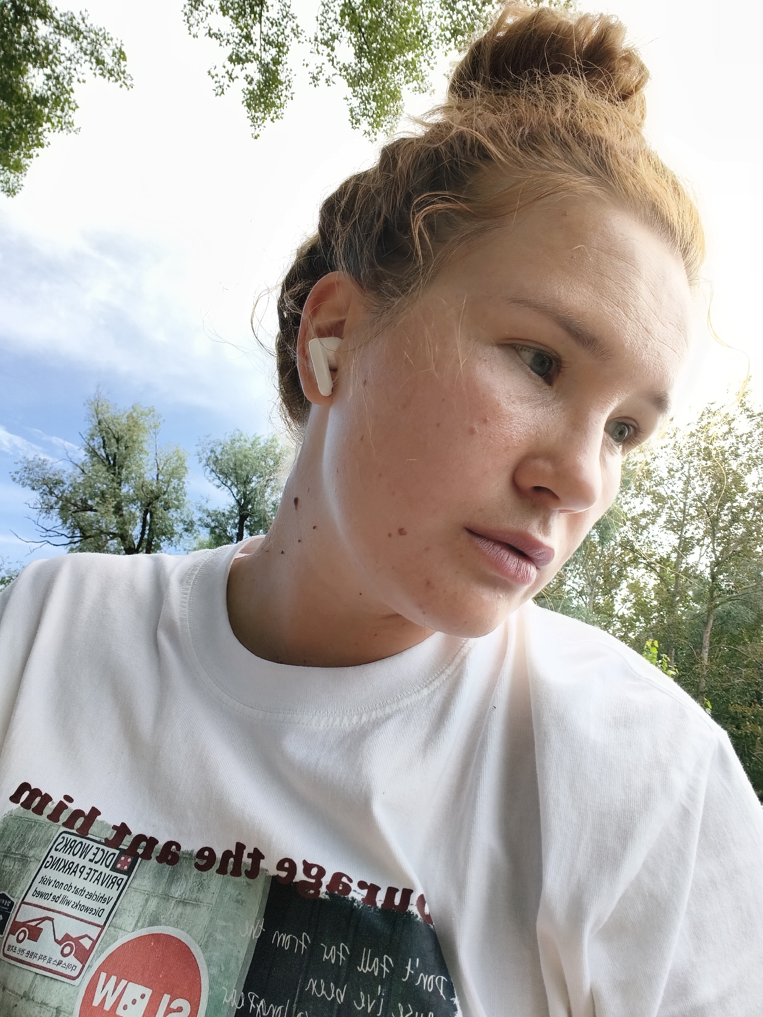 The image shows a close-up, outdoor portrait of a young woman with light skin, reddish-blonde hair pulled up in a bun, and a few freckles. She is wearing a white t-shirt with a graphic print. The graphic on the t-shirt features text and imagery, including a red circle with "W BIA" inside, a sign with "PRIVATE PARKING" and an illustration of a tow truck, and other indecipherable text. She is wearing a white earbud in her right ear. The background consists of trees and a bright sky with some clouds, suggesting a sunny day. The woman is looking off to the right of the frame with a neutral expression. Based on the provided information, the image was taken in Chaikovsky, Russia.