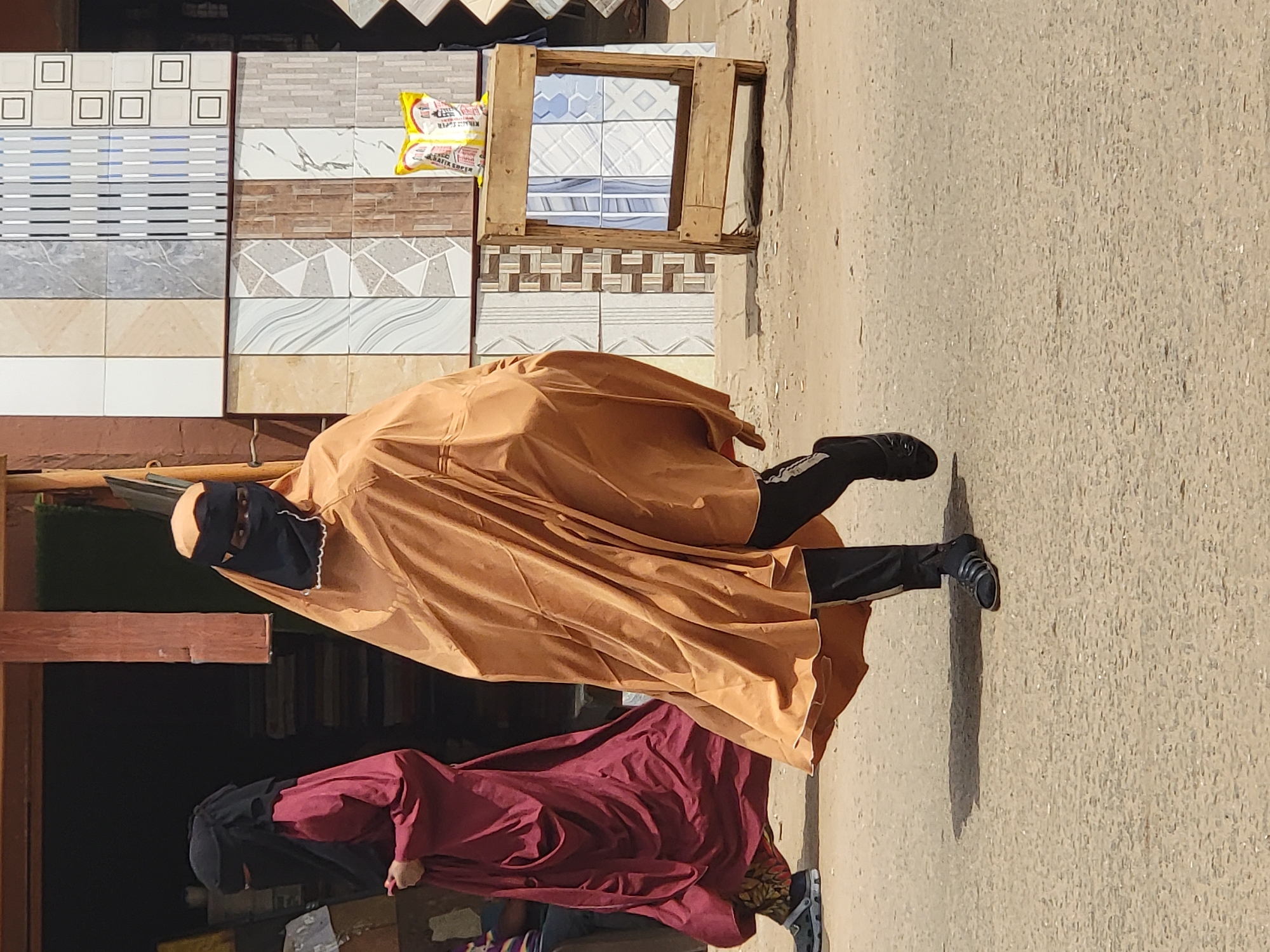 The image depicts two people on a city street in Maiburiji, Nigeria. The individual closer to the camera is wearing a tan-colored cloak-like garment that conceals much of their body, along with black pants and black shoes. They also wear a black face covering. Adjacent to the first individual, a second person is visible, wearing a dark red cloak-like garment. They are positioned further back in the frame and partially obscured. To the left are displays of tiled wall coverings. A wooden frame containing tiles is positioned atop the displays. The street, made of concrete, fills the right of the frame.