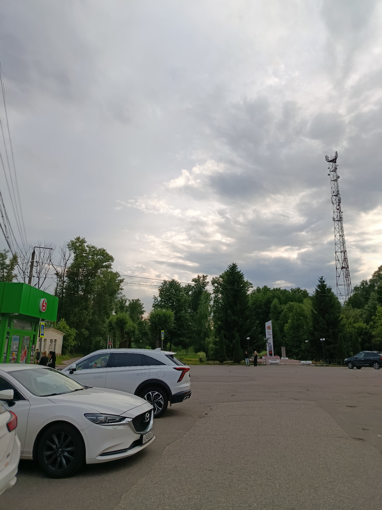 The image depicts an outdoor scene in the city of Vereya, Russia. In the foreground, two white cars are parked on an asphalt surface. The car on the left is a Mazda, and the car on the right is a white SUV. Behind the cars, several trees form a dense line. To the left, a green building with a red circular logo featuring the number "5" is visible, appearing to be a store or gas station. Further in the background, a tall communication tower stands prominently against a cloudy sky. Near the center of the background, a war memorial with inscriptions is present. Several individuals are seen in the distance, near the memorial and the green building. Overhead power lines are strung across the upper left portion of the image. The overall environment suggests an urban or semi-urban setting.