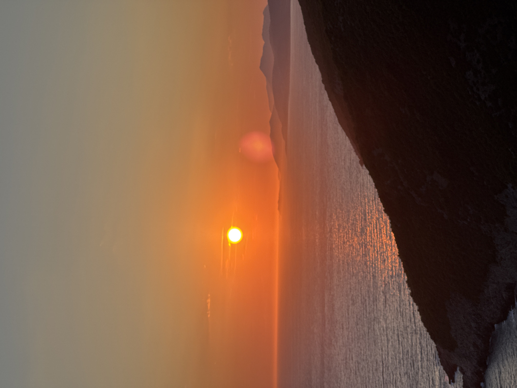 This image captures a serene sunset over the Adriatic Sea, near the city of Zagora, Montenegro. The sun, a bright orb of intense orange, hovers just above the horizon, casting a shimmering golden path across the water. The sea itself is calm, with gentle ripples reflecting the warm hues of the sky. In the foreground, a dark, rugged hillside slopes down towards the water, its texture defined by shadows and subtle variations in light. Distant islands or peninsulas are visible as silhouetted shapes against the colorful sky, adding depth to the landscape. The sky transitions from a vibrant orange near the horizon to a softer, dusky grey at the top of the frame, with a faint hint of pinkish clouds. The overall atmosphere is one of tranquility and natural beauty, characteristic of a Mediterranean evening.
