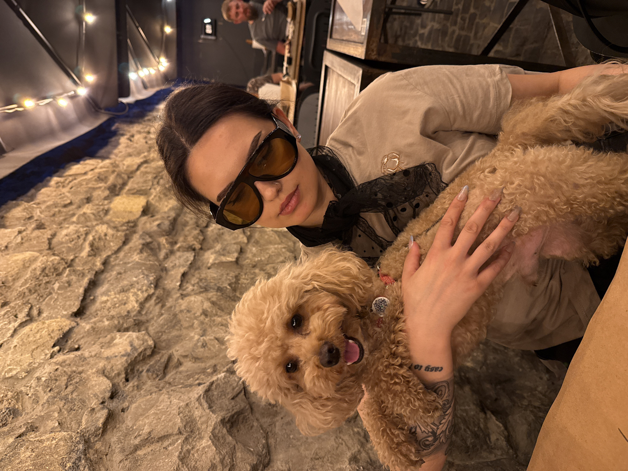 A young woman with dark hair, wearing oversized sunglasses, a beige t-shirt, and a black lace scarf around her neck, is holding a light brown poodle in her arms. The poodle has a red collar and a tag. The woman has a tattoo on her left arm. They are seated at a table with a brown wooden surface in front of them. The background shows a rough stone wall and a ceiling with string lights. In the background, another person is visible sitting at a table, eating. The setting appears to be an indoor restaurant or cafe, possibly with a rustic or industrial aesthetic.