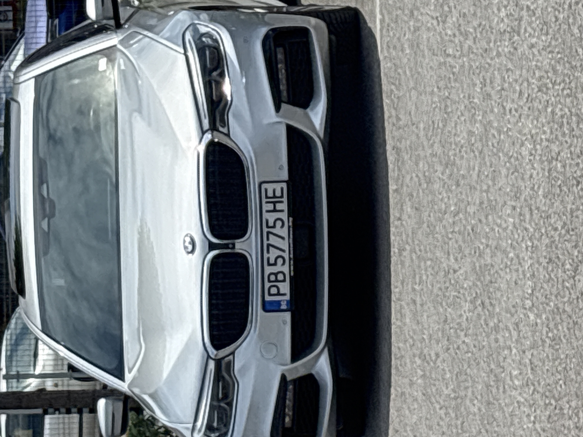 A close-up, eye-level shot depicts the front of a light-colored BMW sedan. The car features a Bulgarian license plate reading "PB 5775 HE." The background consists of a gray concrete sidewalk. Additional details suggest the image might have been taken in an urban environment, possibly Plovdiv, Bulgaria, given the Bulgarian license plate.