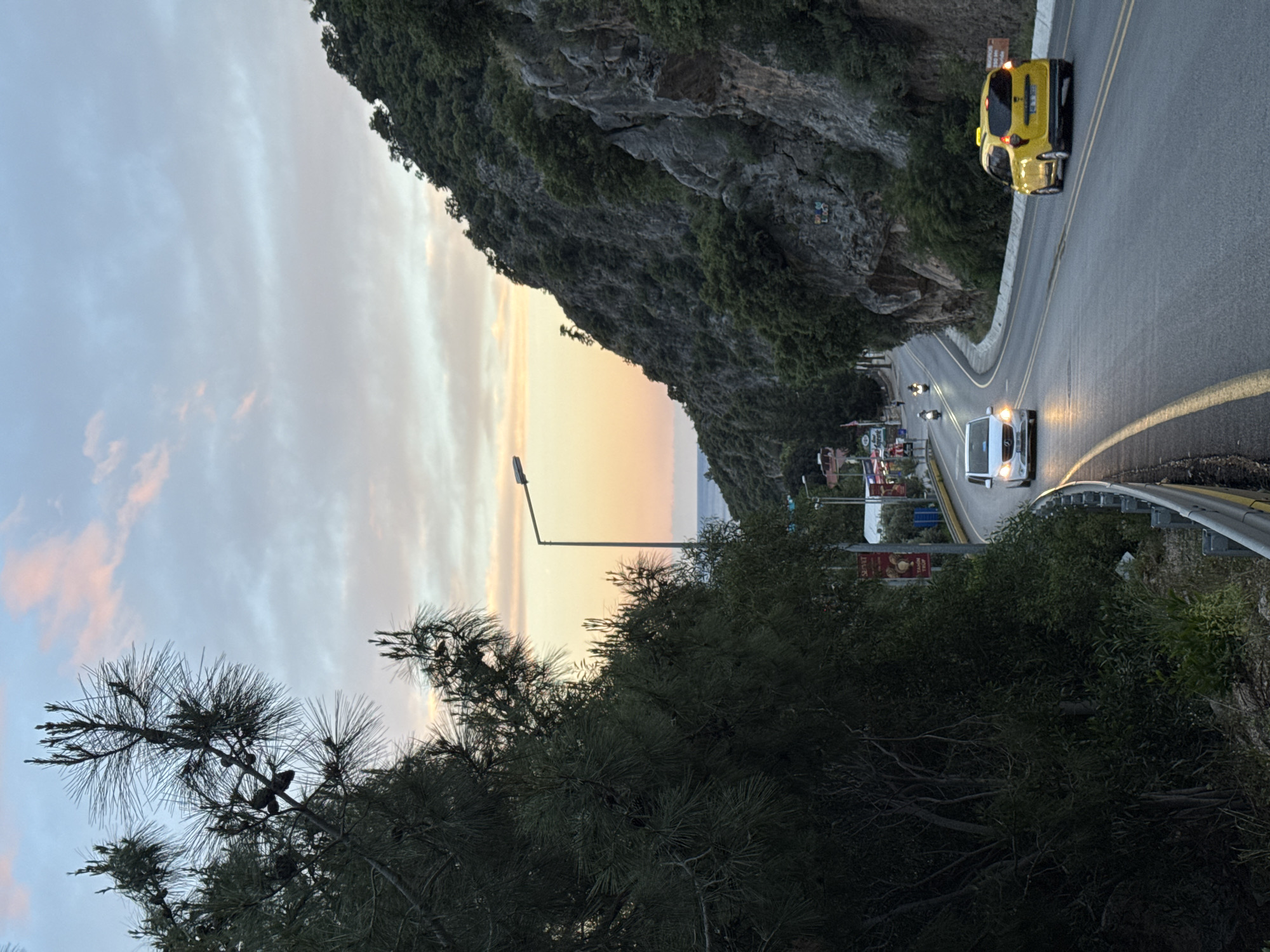 The image depicts a road winding through a landscape near Fethiye, Turkey. A rocky cliff borders the road on one side. Vegetation, including trees, is visible in the foreground and along the roadside. The sky exhibits hues of orange and gray, suggestive of either sunrise or sunset. On the road, there is a yellow taxi and a white car with its headlights on. Street lamps are also present.