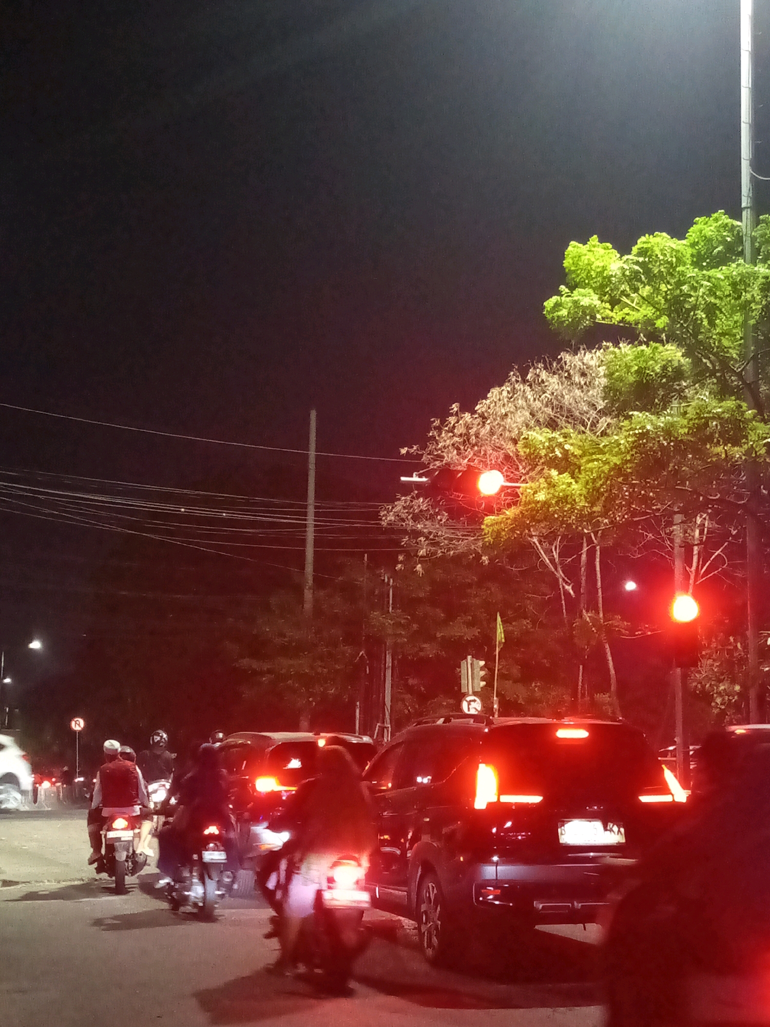 The image depicts a traffic scene at night in Bekasi, Indonesia, likely on a street with a traffic light. Several motorcycles and at least one car are visible. On the motorcycles, multiple individuals are riding, some with helmets, and one appears to be wearing a red vest. The car in the foreground is black and its brake lights are illuminated. Trees with green foliage are present on the right side of the image, with streetlights casting a glow. Power lines are strung across the dark sky. The traffic light is red, indicating that vehicles are stopped. The overall impression is of a typical urban evening commute.