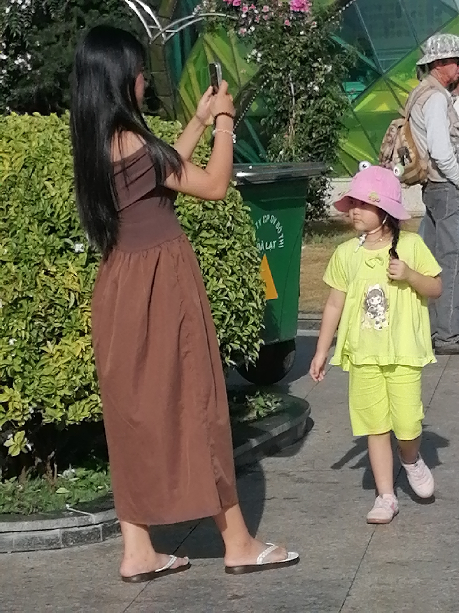 In Da Lat, Vietnam, a woman with long dark hair, wearing a brown off-the-shoulder dress and white flip-flops, stands with her back to the viewer, holding a smartphone up as if taking a picture. Beside her, a young girl in a bright pink bucket hat with frog eyes and a light green t-shirt and shorts, also facing away from the camera, is partially visible. A green trash can with yellow text is to the left, and a blurry background shows lush green foliage and a geodesic dome structure. The scene appears to be outdoors on a paved walkway.