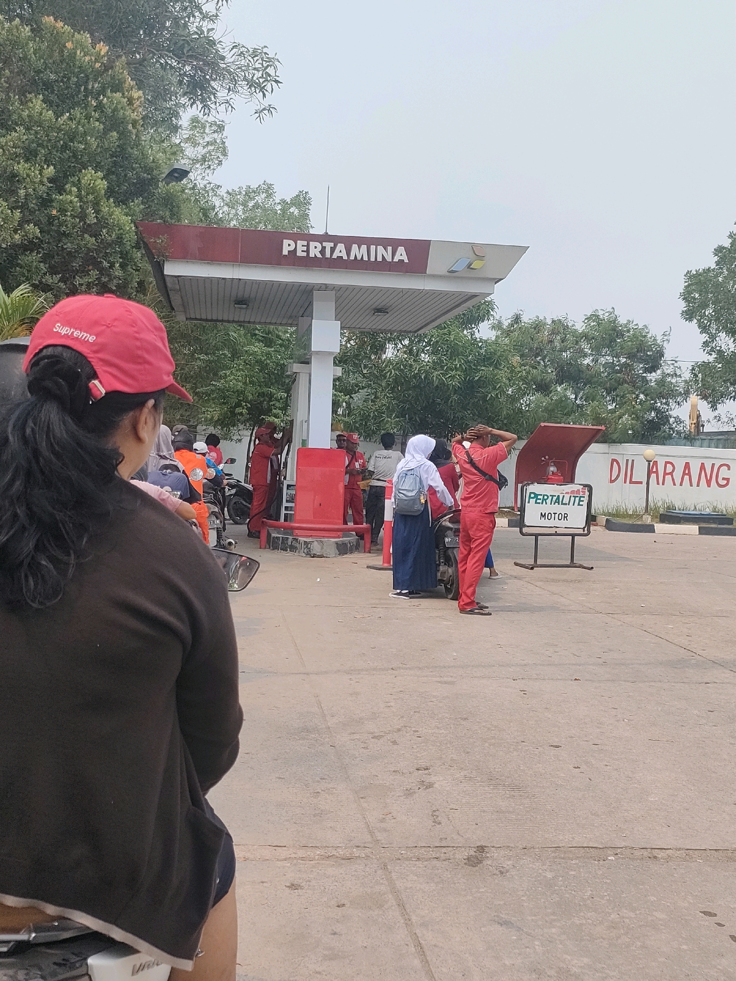 The image depicts a busy Pertamina gas station in Batam, Indonesia. In the foreground, a woman wearing a red "Supreme" cap is seated on a motorbike, facing away from the camera and observing the scene. Behind her, several people are interacting at the gas pumps. Two individuals in red uniforms, likely gas station attendants, are servicing customers. One attendant is seen operating a fuel pump while another stands nearby. Several motorbikes are parked at the station, with people on or near them. A young woman wearing a white hijab and a backpack stands next to a motorbike. In the background, the Pertamina sign is visible on the canopy, along with a prominent red sign that reads "DILARANG" (meaning "Forbidden" or "Prohibited" in Indonesian). The overall environment suggests a typical operational day at a gas station in an urban or suburban area, with the presence of vehicles and customers.