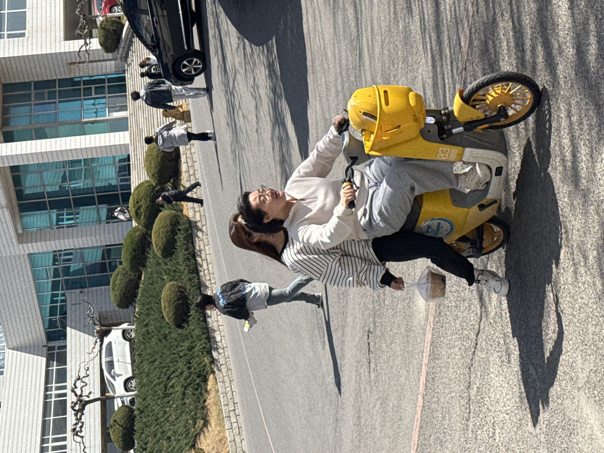 Two young women are riding a yellow scooter down a paved road. The woman driving is wearing a white sweatshirt and gray sweatpants. The passenger is wearing a black and white striped shirt and black pants. A building with glass windows and small, round bushes is on the left side of the road. Pedestrians are walking alongside a black SUV. The location is Zhongtun, China.