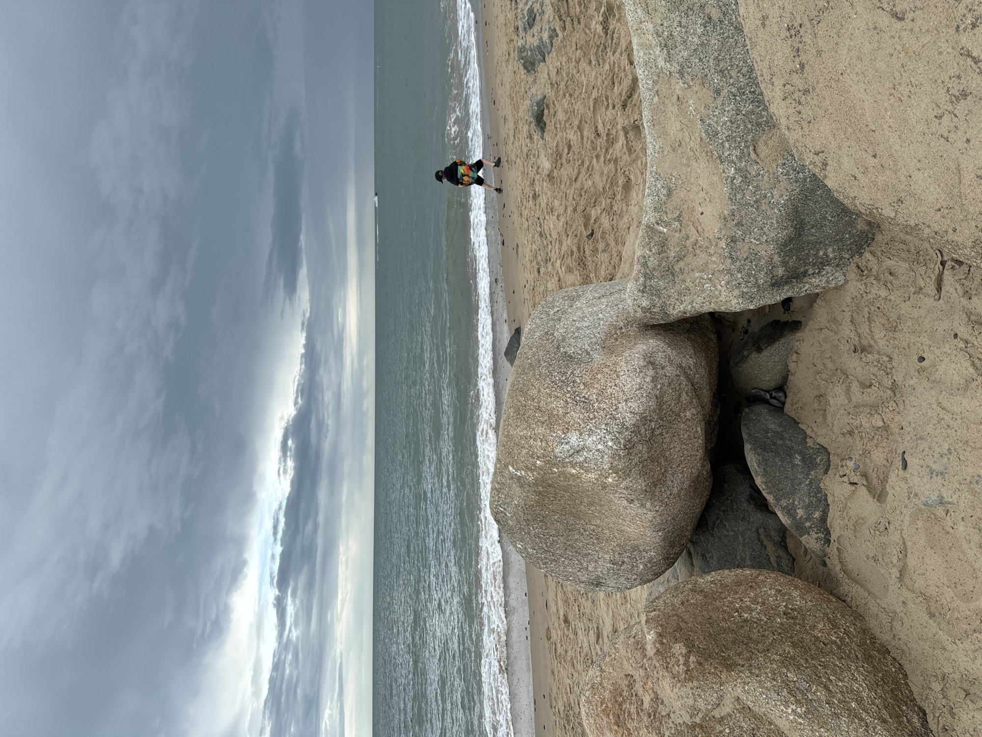 The image depicts a beach scene at Courtown, Ireland, under a heavily clouded sky. In the foreground, large grey and tan boulders are scattered on the sandy shore. The sea is a deep turquoise, with moderate waves breaking on the beach. A person, viewed from behind, wearing a dark cap, a colorful jacket with a tie-dye pattern, and dark shorts, stands on the wet sand near the water's edge, holding a bright orange object. In the distance, a small boat or jet ski is visible on the water. The overall atmosphere is one of a cool, overcast day at the seaside.
