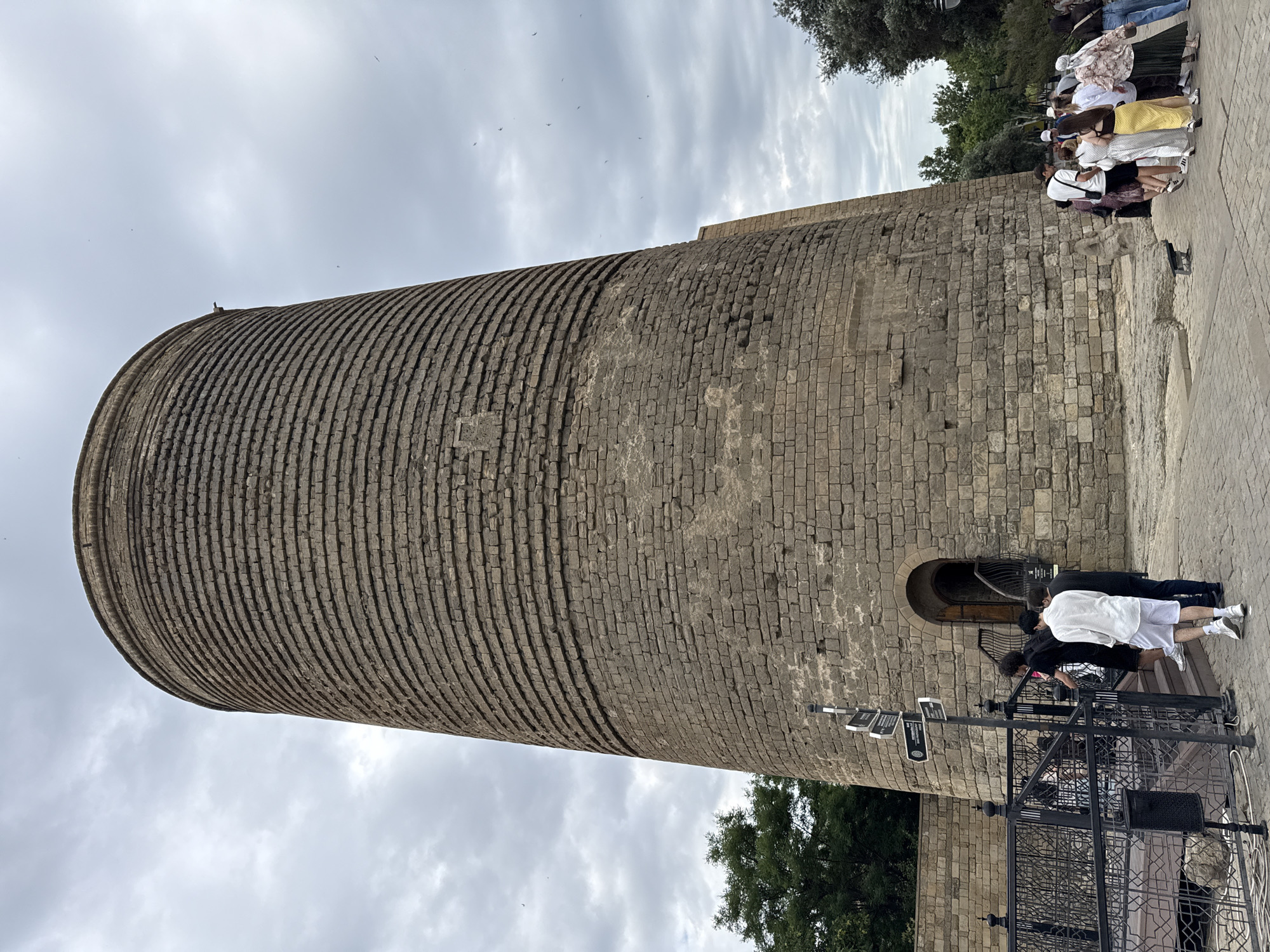 An exterior, low-angle, full shot captures the Maiden Tower, a historical cylindrical stone structure, in Sabail Raion, Azerbaijan. The tower, constructed of rough-hewn stone blocks, exhibits horizontal banding and has a small arched entrance at its base. A group of tourists is visible. On the left, a few individuals are walking up a set of stairs towards the entrance, with a directional signpost nearby. On the right, a larger group of people stands on a paved area, facing away from the tower. Green trees and foliage flank the tower, and the sky above is overcast with clouds and a few small birds are visible in the sky.