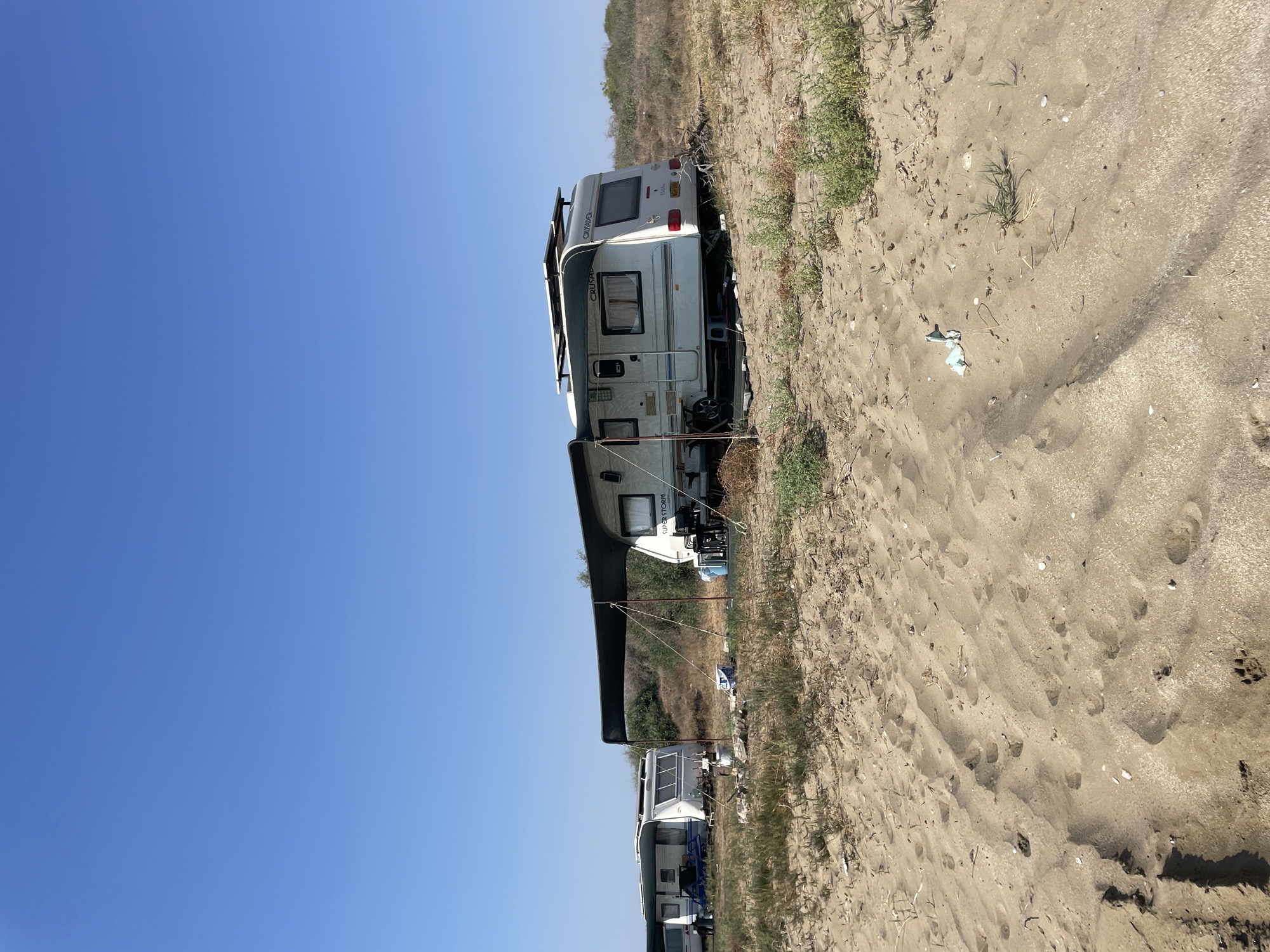 A caravan campsite is pictured in Spathariko, Northern Cyprus. The image shows two white caravans parked on sandy terrain with sparse, dry vegetation. The caravan on the right has an extended awning providing shade over a seating area with chairs and a table. Solar panels are visible on the roof of this caravan. A second caravan is parked to the left of the first, with its own awning extended. The ground is covered in sand with some footprints and scattered debris, including a plastic wrapper. The background features dry bushes and a clear blue sky. No people are visible in the image.