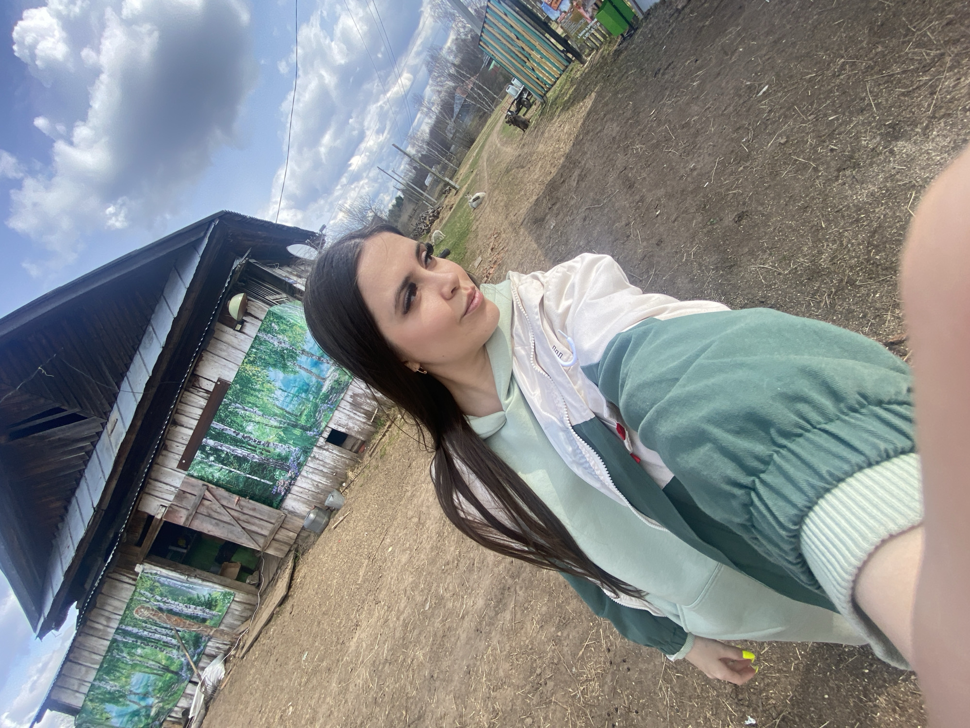 A young woman is taking a selfie outside an old, wooden building in Zhigalki, Russia. The building has landscape paintings on its side and several attached metal buckets. The ground is dirt. In the background, there are trees, a fence, and some wooden structures. The sky is blue with scattered clouds.