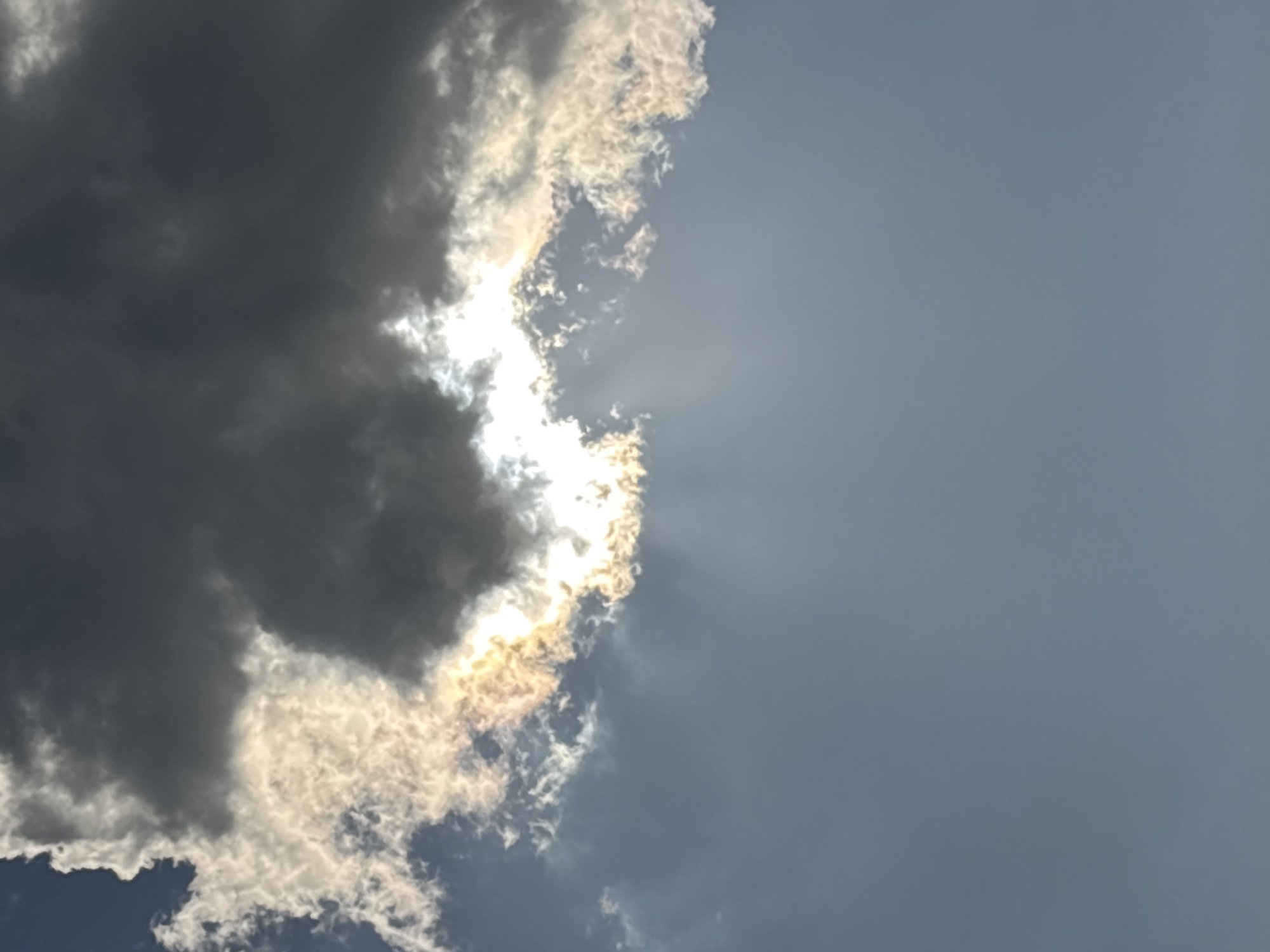 The image is a photograph of a cloudy sky with the sun partially obscured by dark clouds. Rays of sunlight are visible breaking through the clouds, creating a dramatic effect. The lower portion of the image shows a clear, pale blue sky. There are no people, subjects, or objects other than the clouds and sky. The scene does not provide any specific information about the location in the city of Sochi, Russia.