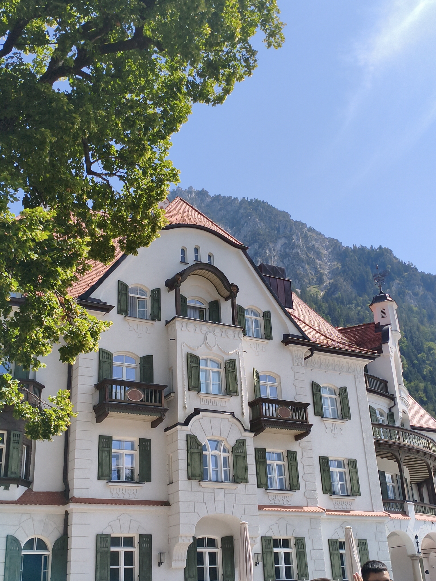 This image captures a picturesque view of a white building with a red tile roof, set against a backdrop of lush green trees and a majestic mountain range. The architecture of the building features multiple stories, ornate balconies with dark wooden railings, and numerous windows with green shutters. The foreground is dominated by the branches of a large tree with vibrant green leaves, partially obscuring the upper portion of the building. The sky above is a clear blue with a hint of sunlight. This scene is consistent with the scenic beauty found in the town of Schwangau, Germany, renowned for its proximity to the Alps and its iconic castles.