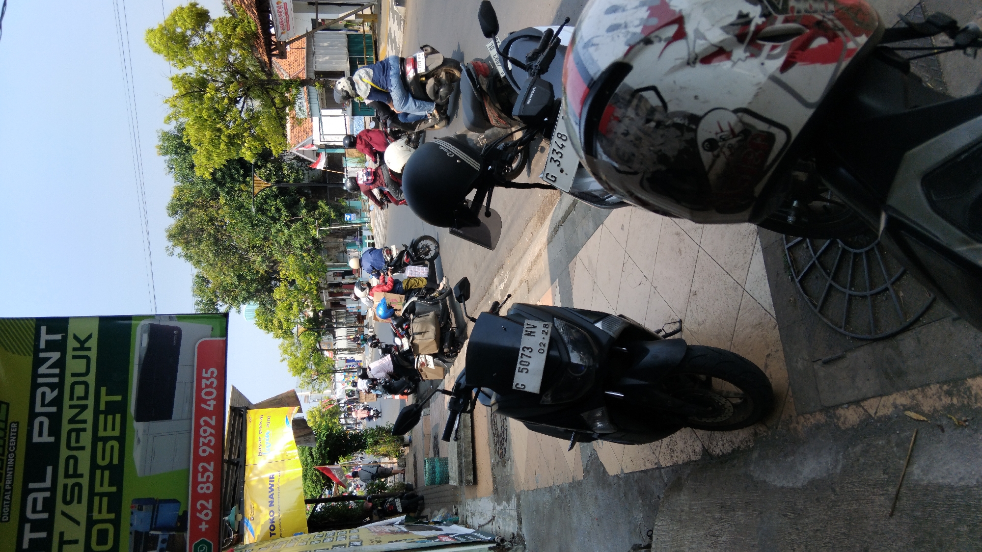 The image depicts a street scene in Batang, Indonesia, with a clear focus on several parked motorcycles and riders in the background. A prominent sign advertising "DIGITAL PRINT T/SPANDUK OFFSET" and a phone number "+62 852 9392 4035" is visible on the left. A yellow banner with "TOKO NAWIR" and "bayar pakai ... aja!" is also present.
In the foreground, two motorcycles are parked, one black and one with a distinctive patterned helmet. The black motorcycle has a license plate "G 5073 NV 02-28". Another motorcycle in the background has a license plate "G 3348".
Several people are seen riding motorcycles in the background. Some are wearing helmets and casual clothing. The environment suggests a typical urban street in Indonesia, with buildings and trees lining the road, and a pedestrian walkway. The sky is clear and bright, indicating daytime. The overall scene conveys a sense of everyday life and commercial activity.