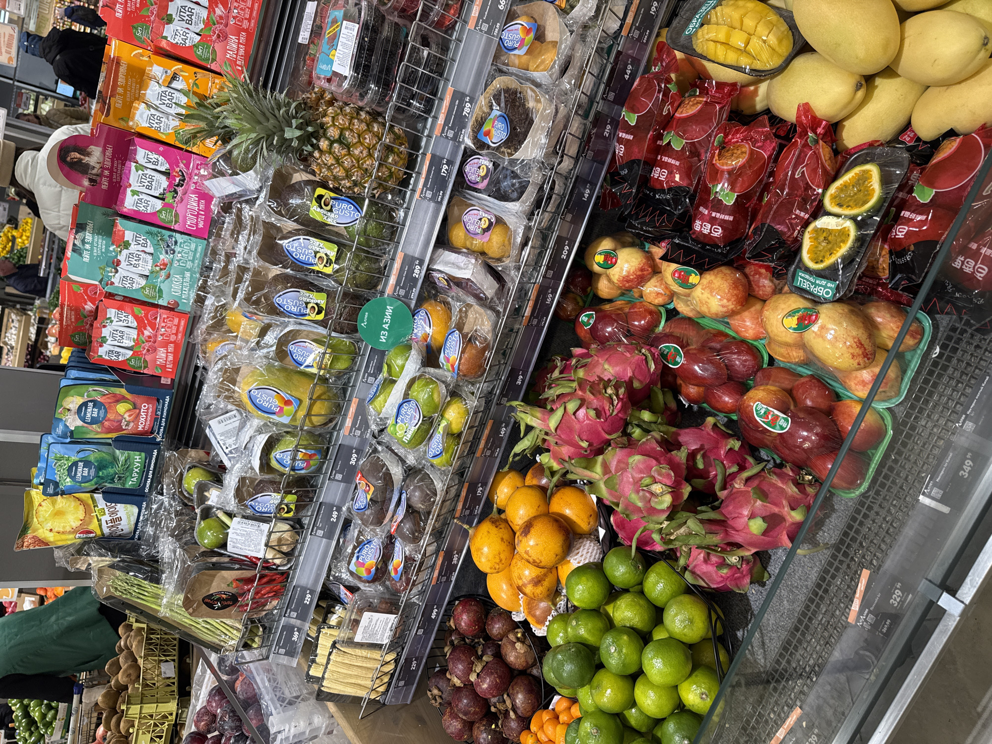 This image captures a vibrant and well-stocked produce section in a supermarket, likely in Krasnoyarsk, Russia, given the contextual information and visible Russian text. The display is brightly lit, showcasing a wide variety of fresh fruits and some packaged goods.
The main subjects are diverse fruits, many of which appear to be exotic. These include large quantities of bright pink dragon fruit, green limes, orange kumquats, and deep purple mangosteens displayed in baskets. On the shelves, there are yellow mangoes, both whole and pre-sliced in plastic trays marked "ОБРАЗЕЦ не для продажи" (SAMPLE not for sale), and numerous red passion fruits, some whole and others cut in half, often pre-packaged. Other visible fruits include avocados labeled "Авокадо Хасс" (Hass Avocado), pineapples, red pomegranates or similar fruits, plums, and various items under the "Puro Gusto" brand, including packaged mini corn and asparagus. An "ИЗ АЗИИ" (FROM ASIA) sticker is visible, indicating the origin of some produce.
Among the packaged items, there are rows of "VITA BAR" fruit bars in different flavors like raspberry, blackcurrant, cranberry & juniper, and thyme & mint, often boasting "Без сахара" (Sugar-free). Above these are "LEMONADE BAR" mixes in flavors like "Мохито Клубничный" (Mojito Strawberry) and "Тархун" (Tarragon), with slogans like "ПЕЙТЕ НА ЗДОРОВЬЕ!" (Drink to your health!) and "ОСНОВА ДЛЯ ЛИМОНАДА" (Base for lemonade).
Two individuals are partially visible: one person in the background wearing a white top and dark jacket, appearing to shop, and another at the bottom left in a green uniform, possibly a store employee. The activity depicted is grocery shopping for fresh produce and related food items.
Notable details include the organized shelving with numerous price tags displaying Russian Ruble amounts, ranging from 66 to 889.99. Some fruit packaging also features Chinese text, such as "百香果" (passion fruit) and "新鲜好品质" (fresh good quality), further indicating the international sourcing of the produce. The overall impression is a modern and diverse supermarket selection.