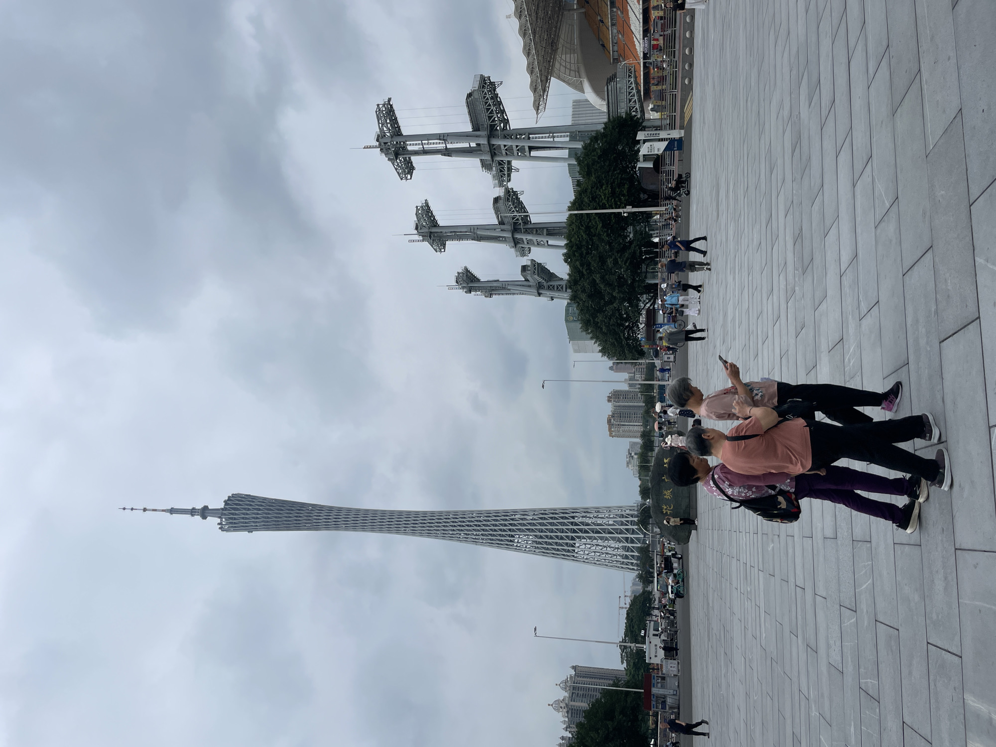 The image captures an outdoor scene in Tianhe District, China, featuring the Canton Tower prominently on the left side of the frame. The tower's lattice structure is visible against a cloudy sky. In the background, multiple large metal structures are seen, possibly related to a stadium or event venue. In the foreground, two women are walking together on a wide, paved area. One wears a peach-colored shirt and black pants, and the other is wearing a purple shirt and purple pants. Other people can be seen walking in the distance. The overall environment appears urban with tall buildings and trees present in the background.