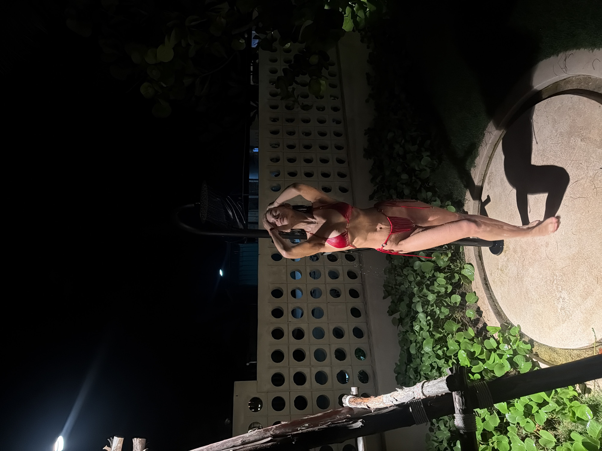 A woman in a red bikini is lying on a bench next to a pool in Choeng Thale, Thailand. The woman is lying on her back with her hands above her head. The pool is surrounded by plants. A concrete wall with circular cutouts is visible in the background. The time is night.