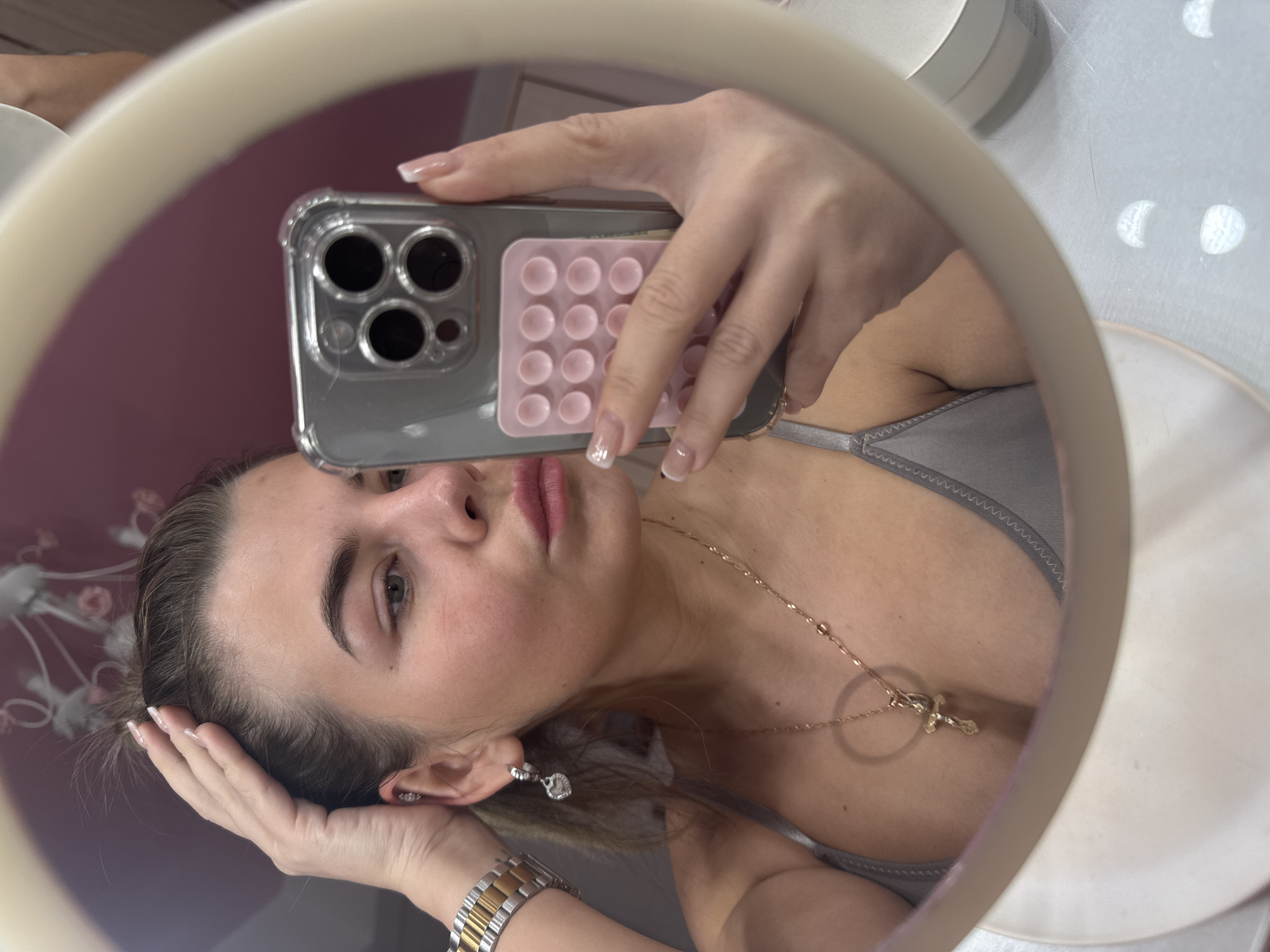 A woman is taking a selfie in a mirror. She is holding a silver smartphone with a pink "pop-it" accessory on the back. The woman has fair skin, brown hair, and manicured nails with white tips. She is wearing a gray top and a gold necklace with a cross pendant. The reflection in the mirror suggests the woman is in a room with a light pink wall and a white table. The presence of tableware items on the table hints to a potential restaurant setting. While the exact location cannot be determined from the image alone, the prompt specifies Chaikovsky, Russia, as the city.