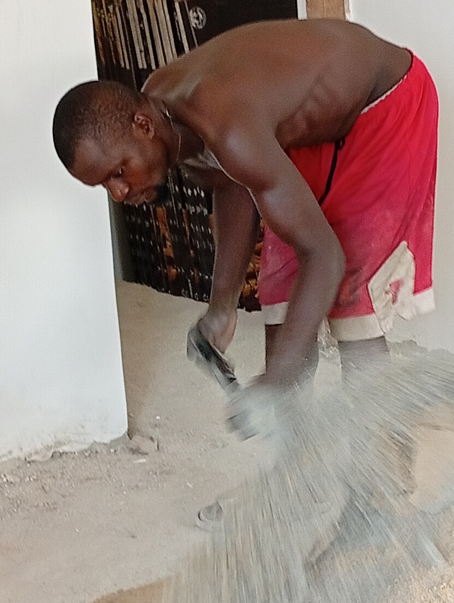 A shirtless man in red shorts is bent over, working on the floor. He appears to be smoothing a substance on the floor with a tool in his hands. Debris or liquid is being splashed out from the action. The location is indoors with a white wall in the background. The city and exact location are unknown.