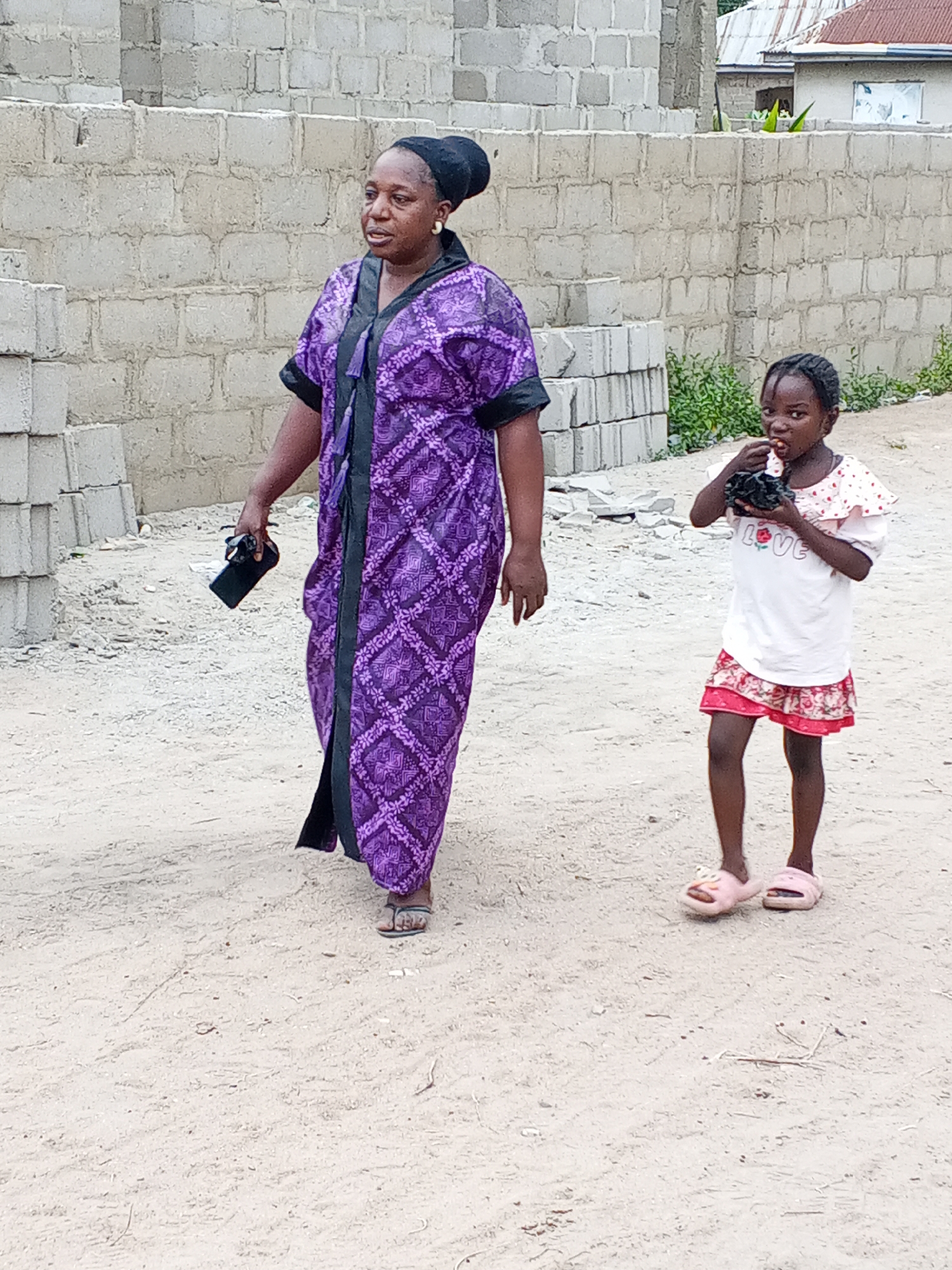 The image captures a woman and a young girl walking on a dirt path in Zango-ndaji, Nigeria, during the daytime.
The woman, on the left, is wearing a long purple patterned kaftan with black trim, a black headwrap, and gold hoop earrings. She is holding a black smartphone in her right hand and wears open-toed sandals. She appears to be walking forward, looking slightly to her right.
The young girl, on the right, has braided hair and is wearing a white short-sleeved t-shirt with "LOVE" written in red letters on the front, and red polka-dot detailing on the shoulders. She has on a red skirt with a floral pattern and pink slide sandals. The girl is holding a small, dark object to her mouth, seemingly eating something, and is looking directly at the viewer.
The setting appears to be a developing or residential area. In the background, there are unfinished walls made of concrete blocks, and stacks of similar blocks are visible on the left. Beyond these structures, sparse green vegetation and parts of simple houses with corrugated metal roofs can be seen. The ground is unpaved, consisting of light-colored dirt or sand. The lighting suggests it is daytime, with soft shadows indicating either an overcast sky or diffused sunlight.