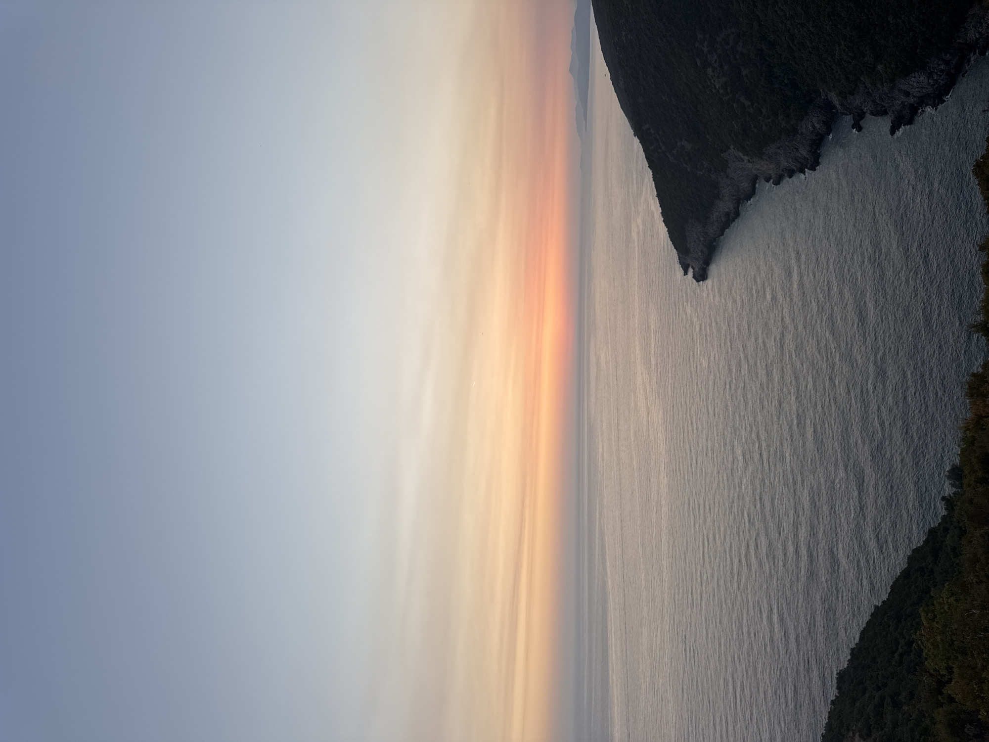 The image captures a serene coastal scene at dusk, likely near Zagora, Montenegro. The sky displays a gradient of colors, transitioning from a pale blue at the top to soft hues of orange and pink closer to the horizon, indicating the setting sun. A vast expanse of calm, gray-blue water fills the lower half of the frame, reflecting the muted sky. On the right side, a dark, tree-covered hill slopes down to the water's edge, forming a natural cove. The coastline is rugged and rocky. On the far right, faint outlines of distant hills are visible against the horizon. The foreground on the bottom left shows the dark, dense foliage of trees on another hill, suggesting a vantage point from a higher elevation. No people, animals, or man-made structures are clearly discernible in the image, emphasizing the natural beauty and tranquility of the location. The overall impression is one of peacefulness and natural solitude.
