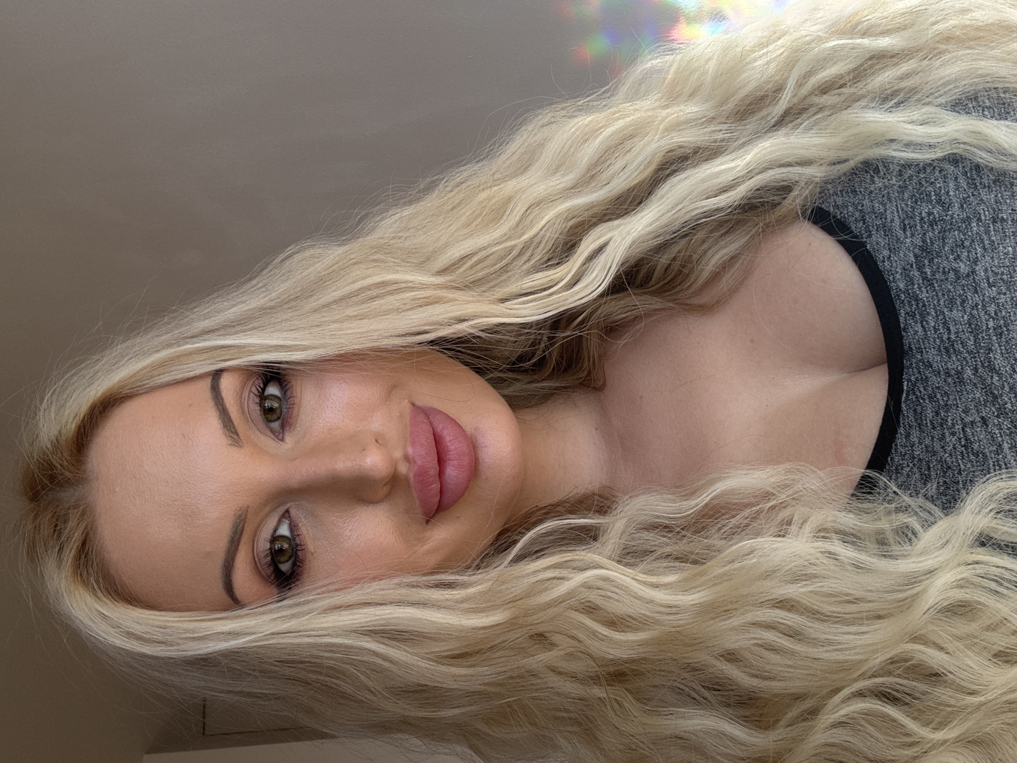A medium shot shows a woman with long, wavy blonde hair and light skin from the chest up. She is looking directly at the camera with green eyes and full, pink lips. Her makeup includes dark eyeliner and mascara, as well as blush and lipstick. She is wearing a grey tank top with a black trim. The background is a plain, light brown wall with a subtle rainbow-colored reflection on the right side of the image.