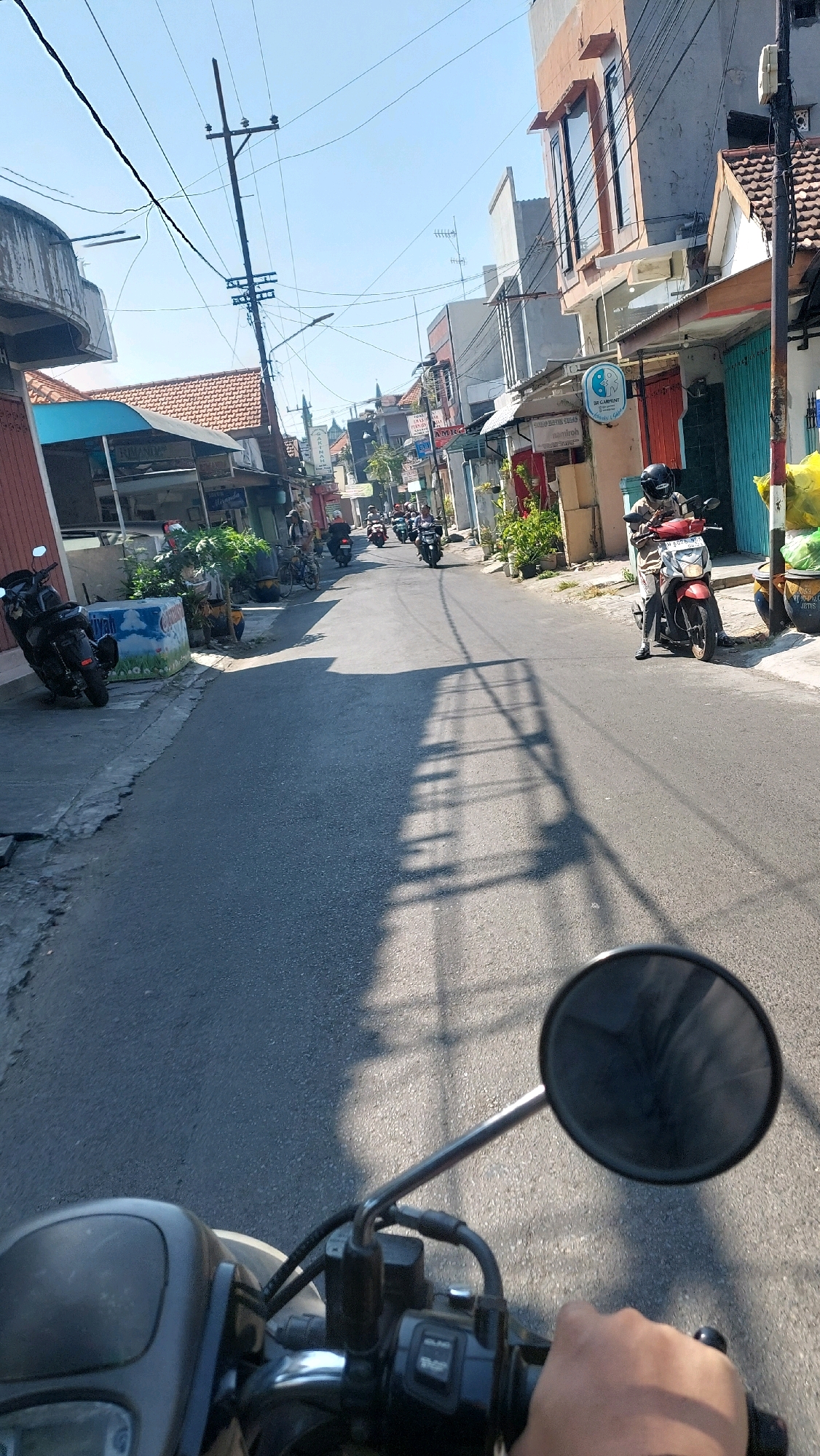 A first-person perspective view of a person riding a motorcycle down a street in Sidoarjo, Indonesia. The street is lined with buildings, some of which are shops or residences. Several other motorcycles are also visible on the road, with riders wearing helmets. Overhead, a network of power lines crisscrosses the sky, supported by utility poles. Shadows of the power lines and nearby trees are cast across the asphalt road, indicating a sunny day. The motorcycle has a black body and a red front panel. The rider's hand is visible on the handlebar, indicating they are operating the vehicle. The overall scene depicts a typical street in a Southeast Asian city, likely during daylight hours.
