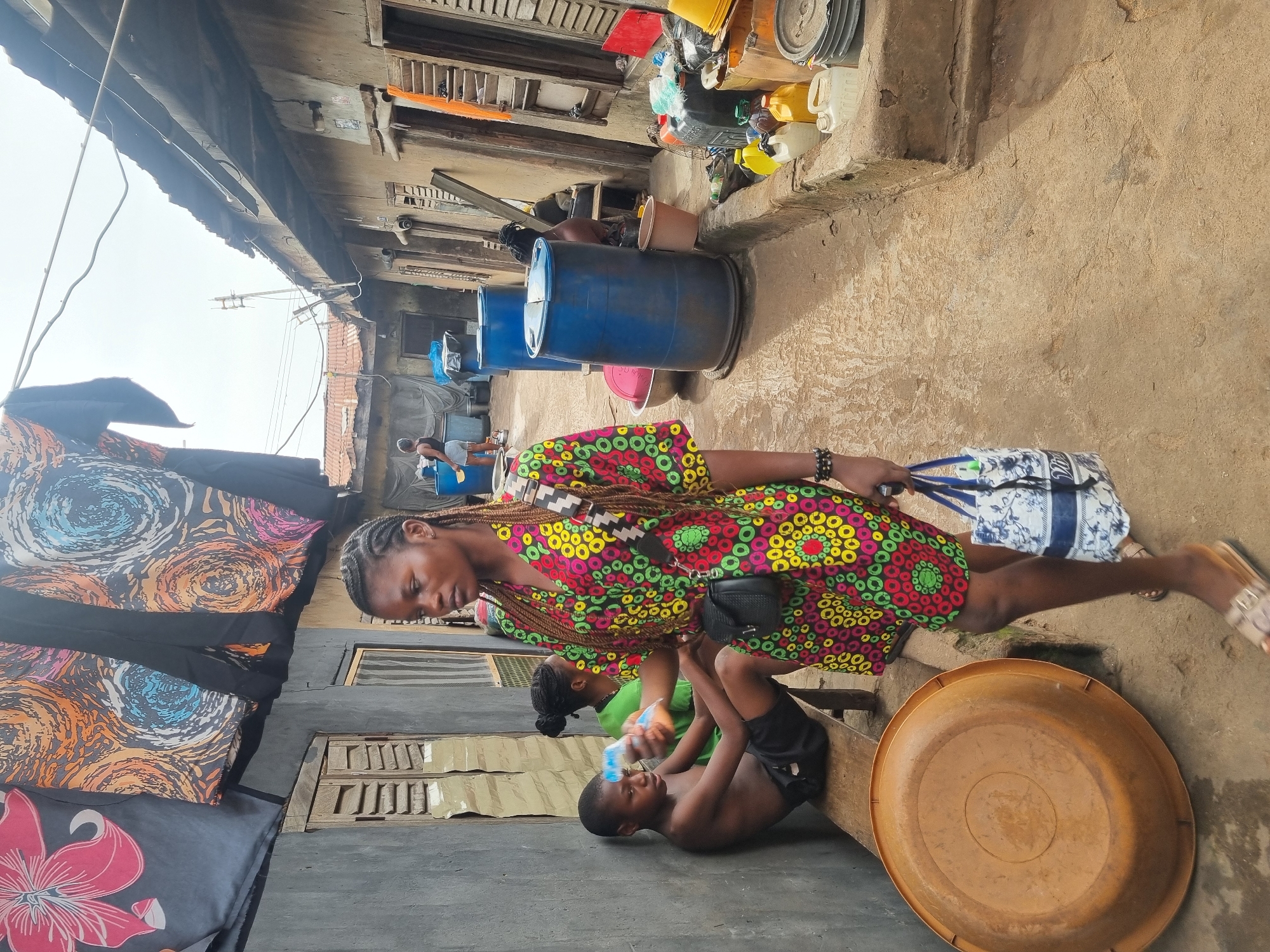 In Onitsha, Nigeria, a narrow alleyway is crowded with buildings and assorted objects. A young woman wearing a colorful patterned dress walks through the alley, carrying a bag. A shirtless boy lies on a wooden plank, and another person sits behind him, appearing to tend to him. Above, laundry hangs from a building. To the right, blue barrels and various containers are stacked against a wall. The ground is textured, suggesting a combination of dirt and concrete. The sky above appears overcast.
