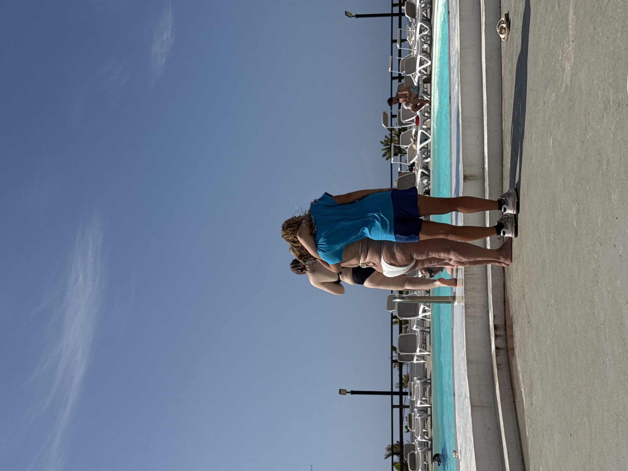 The image, taken in Capaci, Italy, captures a bright, sunny day at an outdoor pool. The sky is a clear, vibrant blue with a few wispy clouds. In the foreground, a person is standing on a concrete patio, wearing a blue t-shirt and navy blue shorts, with their legs and feet visible. They are wearing athletic shoes. Further back, near the edge of a turquoise swimming pool, three individuals are positioned. One person in a blue top and dark bottoms, another in a white bikini, and a third person whose lower body is visible, also in a white bikini. They appear to be standing or perhaps interacting with each other.
The setting includes rows of white lounge chairs, some occupied and some empty, arranged beside the pool. A black fence with vertical supports runs along the side of the pool area, and palm trees are visible in the background, suggesting a warm climate. The pool itself is filled with bright blue water. The overall atmosphere seems relaxed and leisurely, typical of a poolside scene on a sunny day. The angle of the sun suggests it is likely midday. No visible text is present in the image.