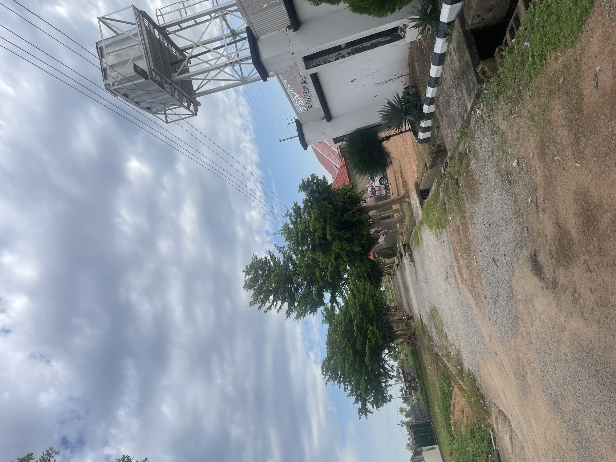 This image depicts a street scene in Jauro Yinu, Nigeria. The street is unpaved, with a gravel and dirt surface. On the right side of the street is a white concrete wall with black and white decorative stripes along its base, topped with a black railing and electric fence. Adjacent to the wall is a large, silver water tower on a metal frame structure, with power lines running across the sky. Behind the wall, parts of a building with a red roof are visible. On the left side of the street, a row of trees with dense green foliage lines the path, casting shadows. Beyond the trees, there are small structures, some with corrugated metal roofs, and green vegetation. The sky is cloudy with patches of blue visible. A person is riding a motorcycle down the street in the distance. A drainage channel runs alongside the road.