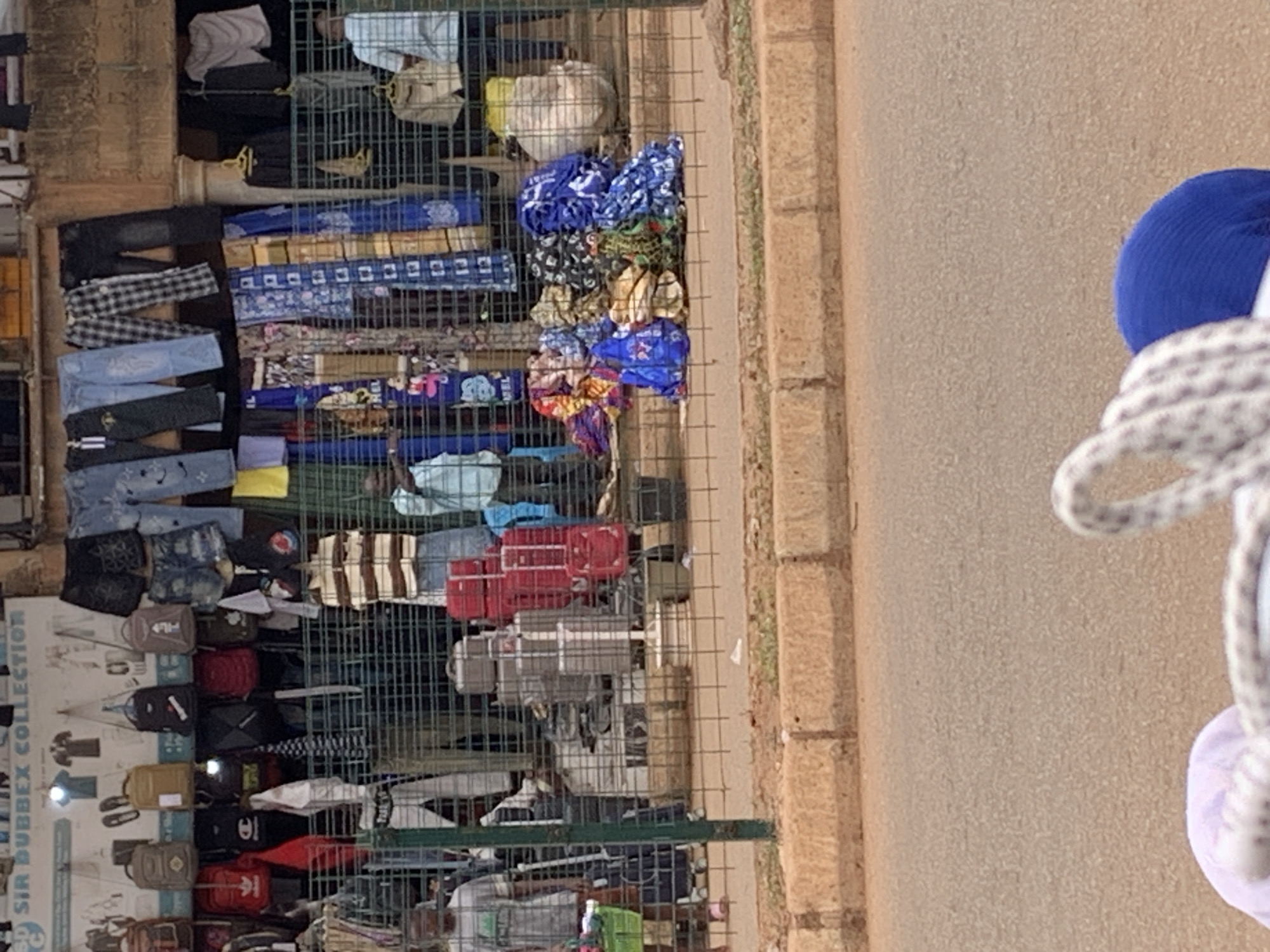 A street market stall is displayed in Bukuru, Nigeria. The stall sells clothing, bags, and suitcases. In the foreground, a pavement is visible, with a street beyond. In the midground, a metal fence encloses the market stall. Behind the fence, various items are displayed, including jeans, t-shirts, bags, and patterned fabrics. Several people are visible behind the fence, some appearing to be vendors and others customers. One person in the foreground is out of focus. In the background, buildings and a sign reading "SIR DUBBEX COLLECTION" are visible.