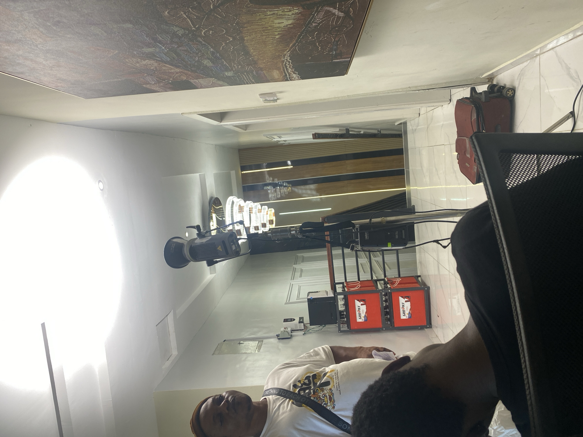 This image captures an indoor setting, likely a production area or modern event space, filled with specialized equipment and contemporary decor. The original photo is rotated 90 degrees counter-clockwise; described here as if correctly oriented.
In the foreground, to the right, only the back of a person's head and shoulder are visible, wearing a dark top, with a black office chair beside them. In the mid-ground, slightly to the left, another person is seen from the chest up, wearing a white t-shirt with a graphic design that includes partial text like "SOMETHING TO EACH OT..." and a sling bag across their chest. They are also wearing a light-colored cap or head covering.
The space is dominated by professional equipment. On the left, a very bright, artificial light source emits a strong glare. Further into the mid-ground, a professional video or film light, possibly branded "LUPHEN," is mounted on a stand. Adjacent to this, a rack holds several orange-colored battery units clearly labeled "GROVOLT," alongside a smaller black box, indicating a power supply or backup system.
The background reveals a modern interior with a long perspective, resembling a hallway or elongated room. The walls feature decorative paneling with integrated horizontal lighting. A red rolling suitcase is leaning against a tiled wall on the far right. The ceiling is a notable detail, featuring a large, framed piece of dark, intricate artwork or tapestry, alongside recessed and linear lighting elements.
The scene suggests activity related to media production, a photoshoot, or an event setup requiring dedicated lighting and power. The intense artificial lighting is a prominent feature, making the time of day indistinguishable.