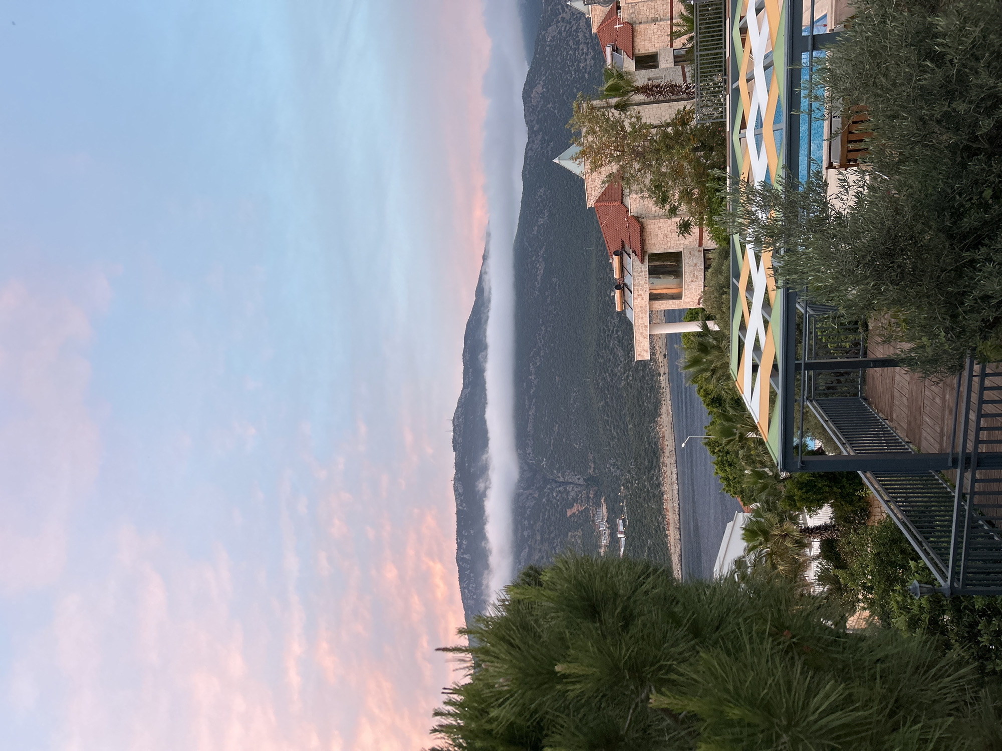 The image depicts a scene in Kaş, Turkey. It shows a mountain range partially obscured by a band of low-lying clouds or fog. The sky is a gradient of pink and blue hues, suggesting dawn or dusk. Buildings with red-tiled roofs are visible on the right side of the image. A structure with a colorful, geometric design overlooks what appears to be a pool area. The foreground includes trees and vegetation. A road runs alongside the mountain's base.