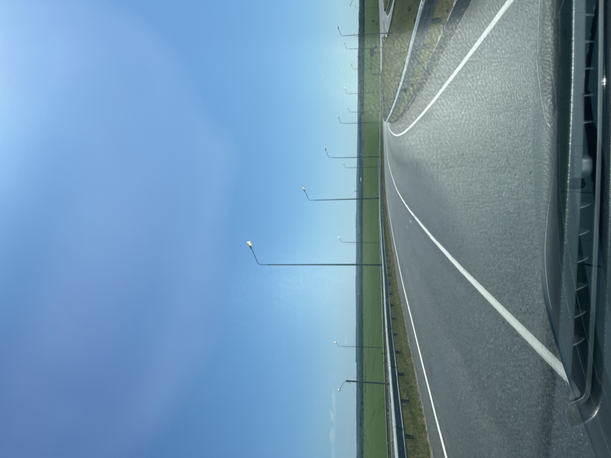 The image depicts a highway stretching into the distance under a clear blue sky, suggesting a bright and sunny day. The perspective is from a vehicle, with the right side of the image showing the car's bodywork, possibly a window frame and part of the car's interior. The highway is paved with asphalt and has distinct white lane markings. Along the left side of the highway, a series of tall lampposts are visible, spaced evenly along the road. Beyond the lampposts, a wide expanse of green fields can be seen, hinting at a rural or semi-rural location. The fields appear flat and stretch towards the horizon, where they meet the sky. The lighting suggests it is daytime, likely late morning or early afternoon, given the clear sky and the intensity of the sunlight. There are no people visible in the image. The overall scene conveys a sense of travel and open road, with the ordered lampposts and clear sky contributing to a feeling of calm and progression. The location context suggests this is a road in Nikol'skoe, Russia.
