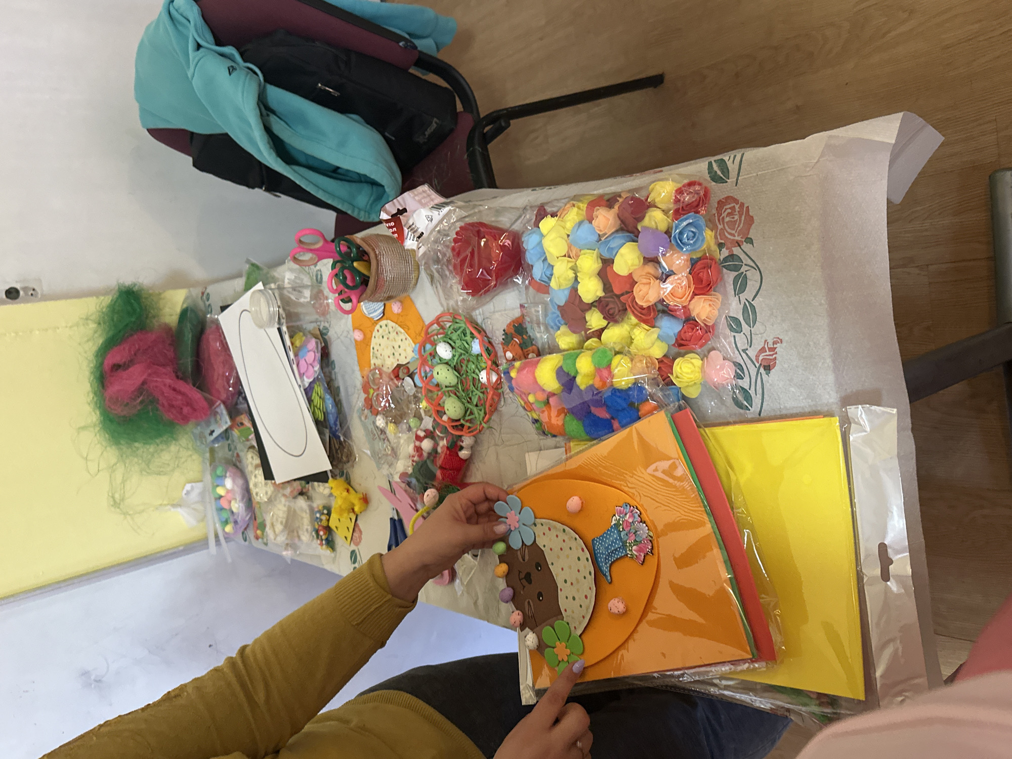 A high-angle shot shows a woman working at a table filled with craft supplies. The scene appears to be indoors. A table covered with a patterned fabric is laden with materials including colored paper, fake flowers, fuzzy balls, and various other embellishments, suggesting a crafting project is underway. A woman in a mustard-colored sweater is holding a craft project with a bunny cutout. The backdrop includes a yellow wall, a chair with a jacket and bag, and a wood floor. No specific indicators point to Barakovo, Bulgaria.