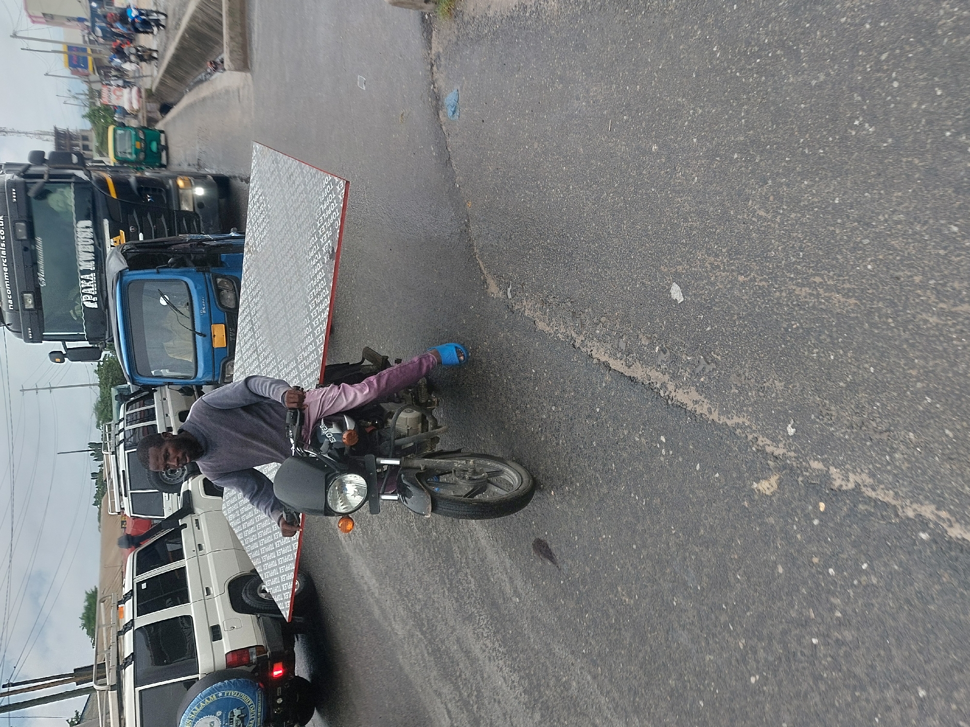A man on a motorcycle is transporting a large, rectangular object covered in text. The text appears to say "TOPPLEX". He is wearing a gray top and pink pants with blue sandals. The motorcycle is black. The scene is on a street, with vehicles, including trucks and a white SUV, visible in the background. The location may be Dar es-Salaam, Tanzania, as some vehicles have East African plates.
