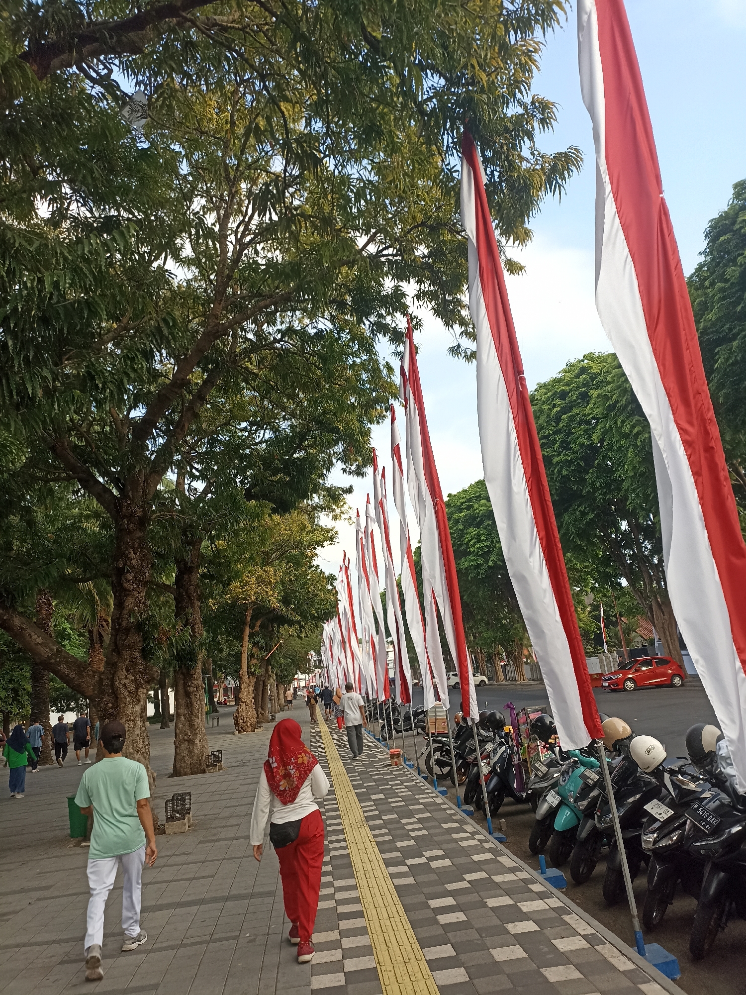 The image depicts a sidewalk lined with Indonesian flags. In the foreground, two individuals are walking away from the camera, one wearing a light green t-shirt and white pants, the other a red hijab and red pants. To the right, a row of parked motorcycles is visible, and beyond them, a red car is parked. Trees with dense green foliage line the left side of the sidewalk, casting shadows on the ground. The sky is clear blue with a hint of sunlight. The scene appears to be a public space within Kelurahan Kepatihan, Indonesia, likely a promenade or walkway, given the presence of pedestrians, flags, and parked vehicles.