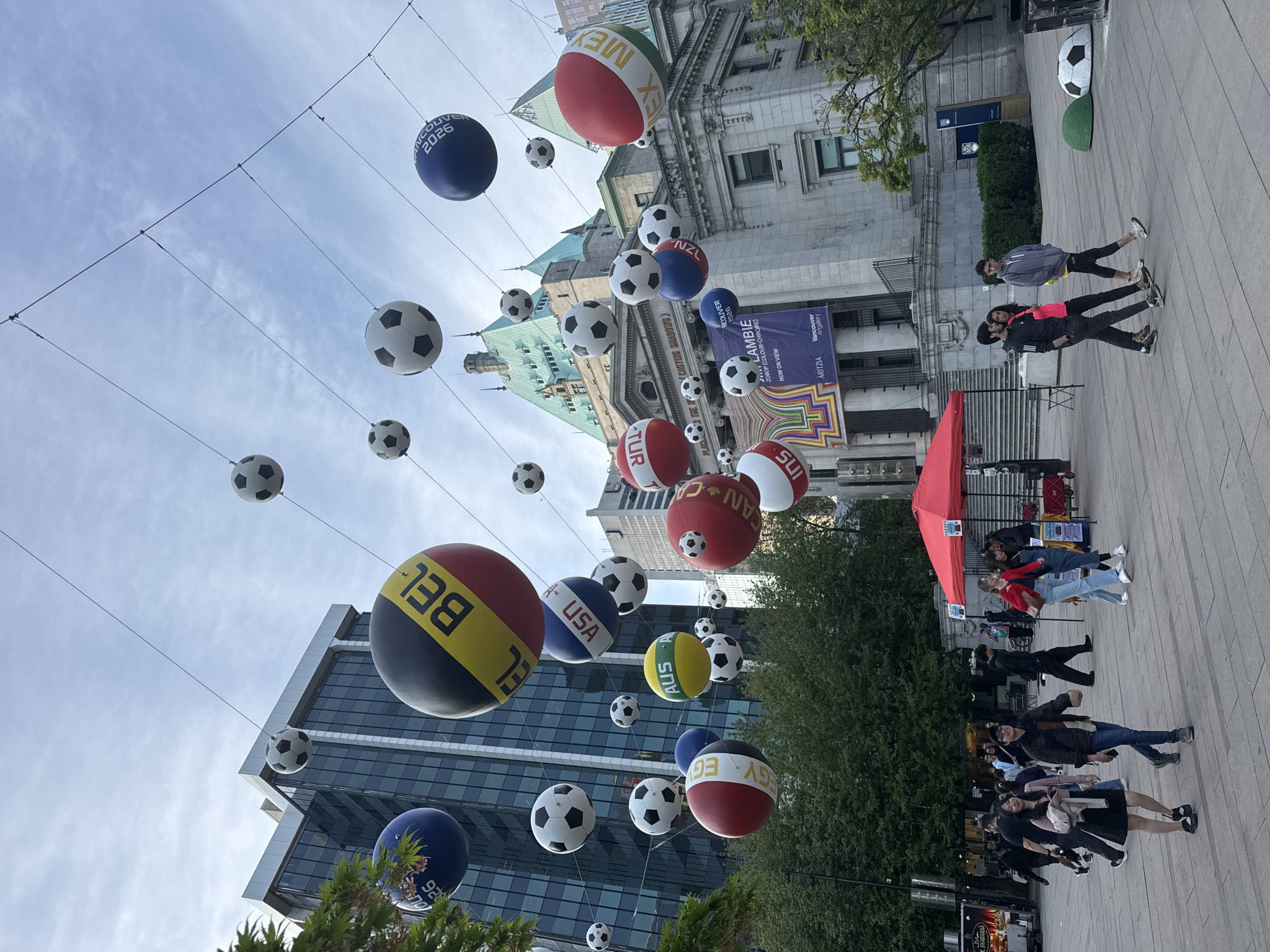 The image shows a street in Vancouver, Canada, decorated with soccer balls of various sizes hanging from wires overhead. The balls are decorated with national flags and the words "Vancouver 2026." The Vancouver Art Gallery building is visible in the background. People are walking on the sidewalk in the foreground, and there is a red tent set up. Buildings surround the street, and the sky is partly cloudy.