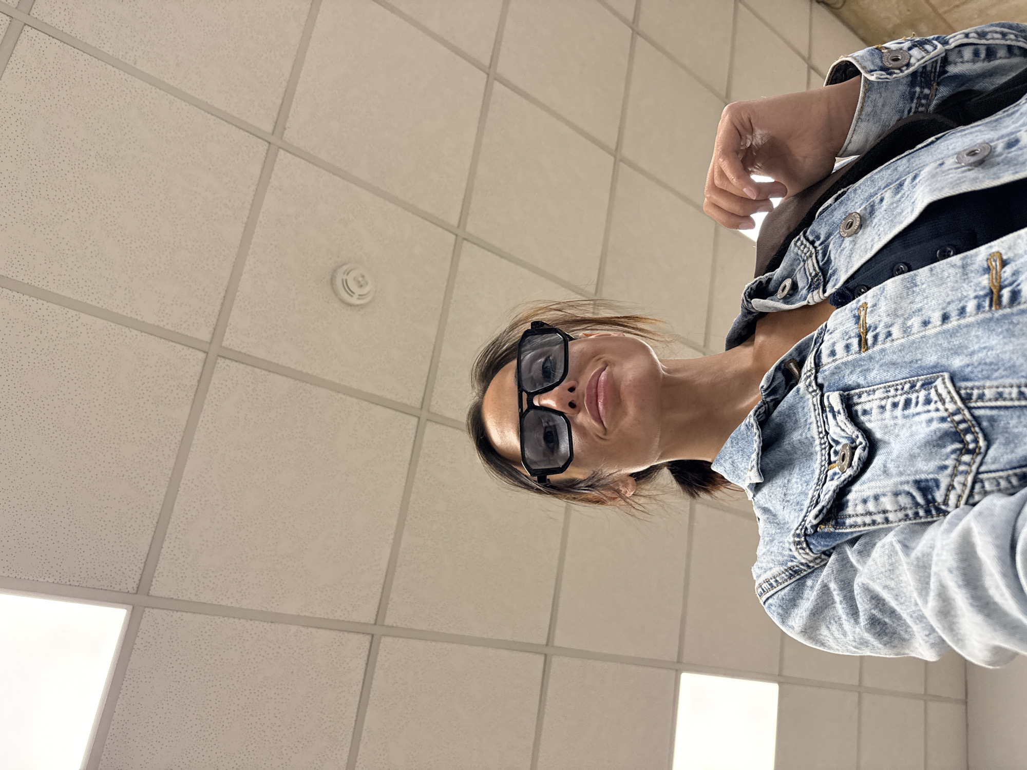 A woman with dark hair pulled back in a ponytail is wearing oversized black sunglasses and a light blue denim jacket over a dark top. She is holding the lapel of her jacket with her right hand, which is resting on her shoulder. Her left hand is not visible in the frame. The background is a dropped ceiling with fluorescent lighting panels and a smoke detector visible. The low angle of the shot suggests the photo was taken by the woman herself, possibly in a public indoor space such as an airport terminal or a shopping mall. Based on the provided metadata, the location is identified as Sofia, Bulgaria.