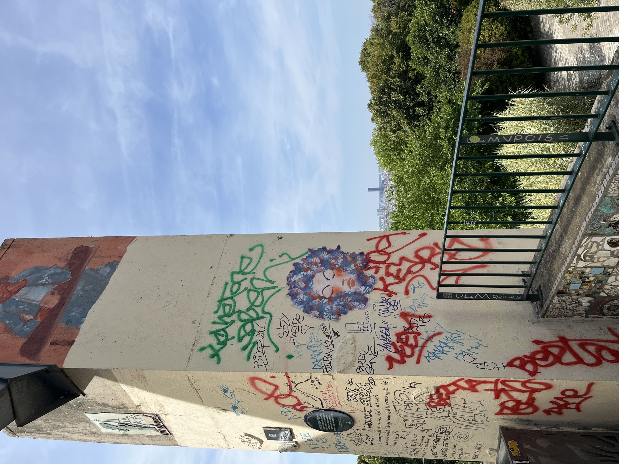The image shows a graffiti-covered tower with a mural of a person in the upper section. The tower is located in Paris, France, with a view of the city skyline and lush green trees in the background under a partly cloudy sky. The tower's surface is covered with various tags and drawings, including a stylized face with curly hair. A metal railing with vertical bars runs along the foreground, separating the tower from a paved area and vegetation. The overall scene is an outdoor urban environment with a focus on street art and city landscape.