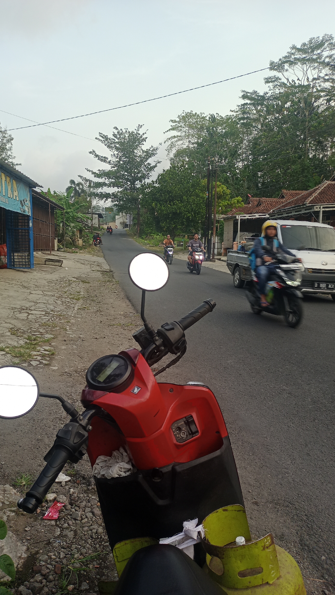 A red scooter is parked on the side of a paved road in Karyamandala, Indonesia. The scooter is in the foreground, and its handlebars, mirrors, and front fairing are visible. A yellow propane tank is attached to the side of the scooter. The road is lined with trees and buildings. Several people are riding motorcycles and a white van is driving on the road. The sky is overcast.