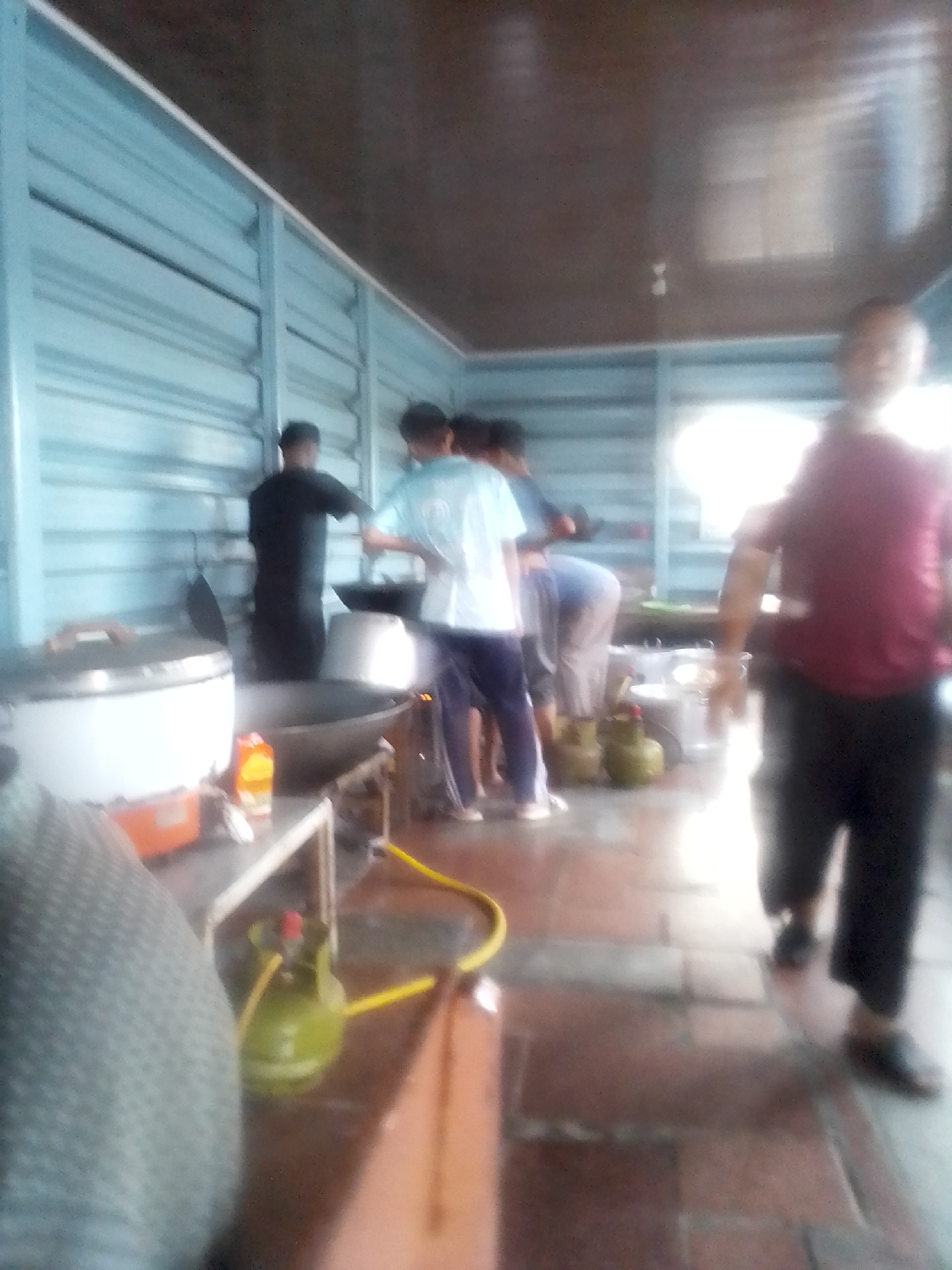 The image depicts an indoor scene, possibly a kitchen or food preparation area, located in Sukasari, Indonesia. The environment appears to be an open-air structure with light blue corrugated metal walls and a dark ceiling. In the foreground, a large white rice cooker with a wooden handle is visible on a table, alongside an orange box and a carton of liquid. To the right, there is a large black wok and a gas stove. Several people are present in the midground, with at least three individuals actively engaged in cooking or preparation. One person in a dark t-shirt is standing by the wok, and another in a light blue t-shirt is also near the cooking area. A third person is bent over a gas tank on the right side of the image. Two green gas cylinders with yellow hoses are also visible on the floor. In the background, a person in a maroon t-shirt and black pants is walking away from the camera. The overall impression is one of a busy food preparation environment.