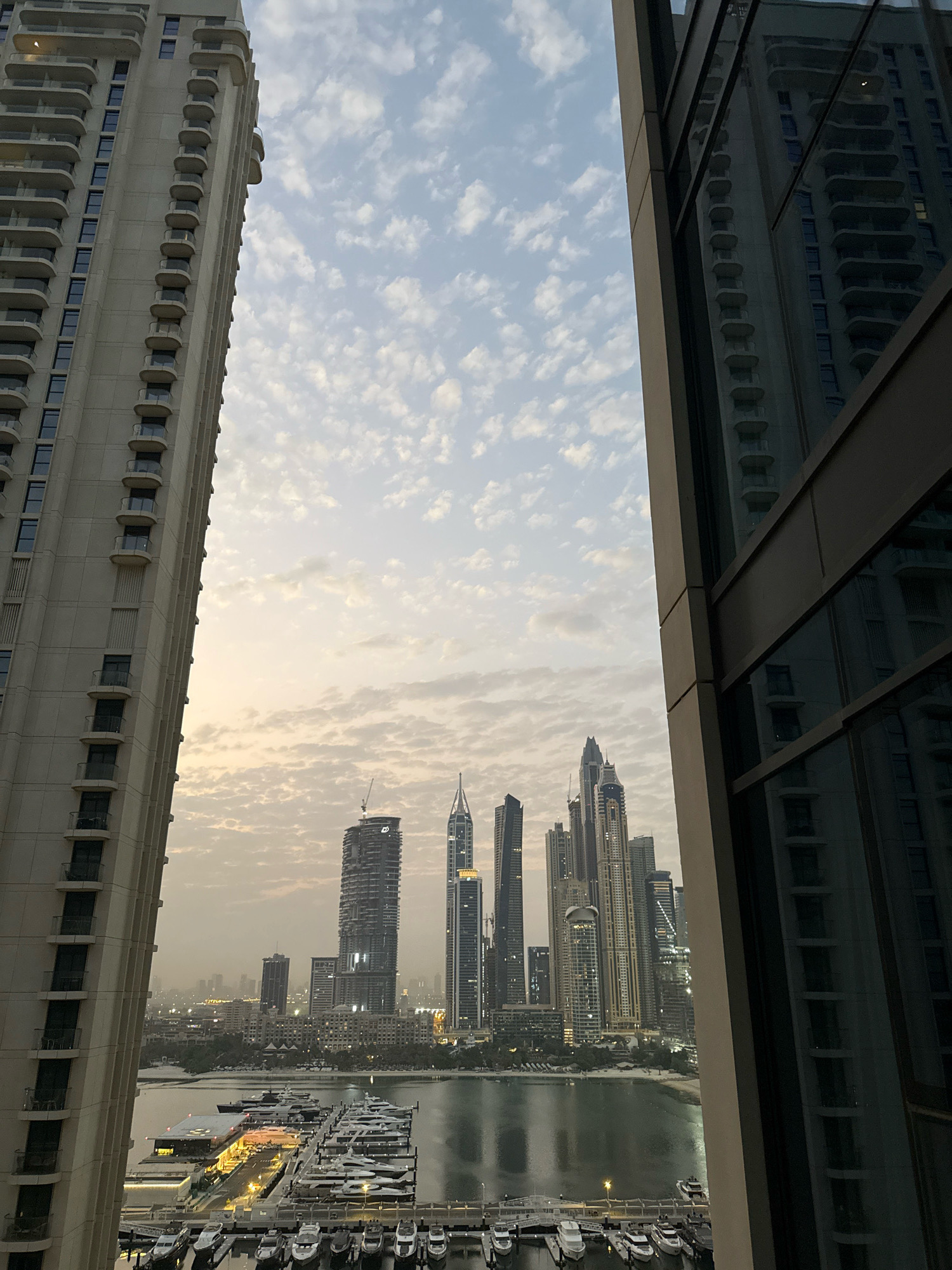 This vertical, eye-level, medium shot captures a view of the Dubai Marina, United Arab Emirates, from a high vantage point. On the left, a tall, modern beige residential building with a repetitive pattern of balconies dominates the frame. On the right, a dark, contemporary building with reflective glass surfaces is partially visible, mirroring the building on the left and the sky.
Between these two structures, the Dubai Marina skyline unfolds. A body of water, likely the marina itself, occupies the lower portion of the scene, filled with numerous docked yachts of varying sizes. Some yachts are brightly lit, suggesting it's either early morning or evening. A long pier or walkway with lights runs through the center of the docked boats.
Beyond the water, the iconic skyscrapers of Dubai Marina rise, showcasing a variety of architectural styles and heights. One prominent skyscraper in the center has a crane at its apex, indicating ongoing construction. The sky above is filled with a scattered pattern of white, wispy clouds against a pale blue and orange gradient, indicative of sunrise or sunset. The overall atmosphere is serene and urban. No people are visible in the scene.