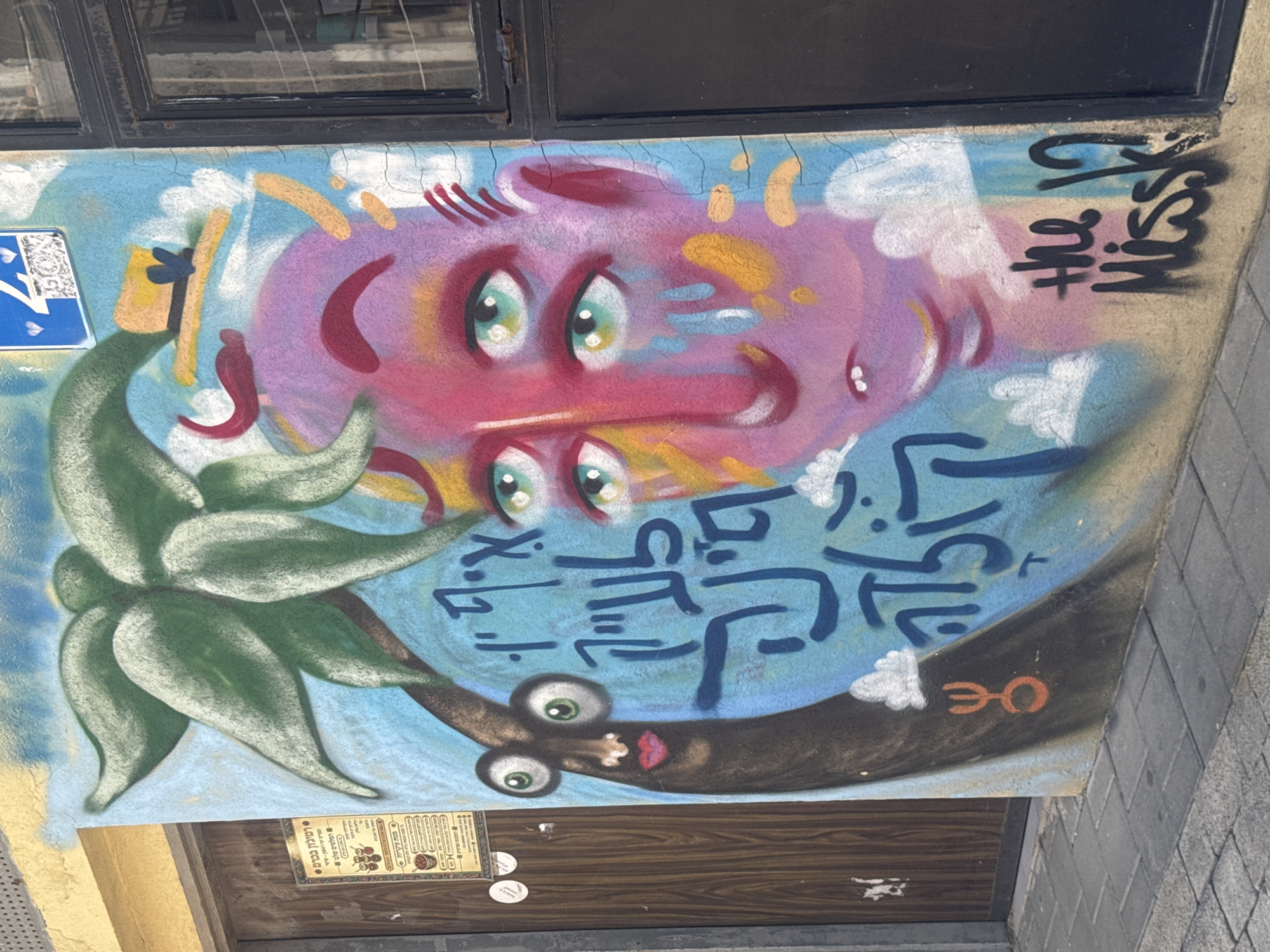 The image shows a piece of street art painted on a wall or concrete surface in Tel Aviv, Israel. The mural includes cartoonish depictions of two faces: one in pink and one in brown, along with Hebrew text. A green plant-like form is painted adjacent to the brown face. The style suggests a somewhat whimsical, urban art aesthetic. The bottom of the image shows what appears to be a doorway or entrance, with a sign on it, indicating the location is in Tel Aviv. The Hebrew text and overall artistic style suggest a local Israeli influence.