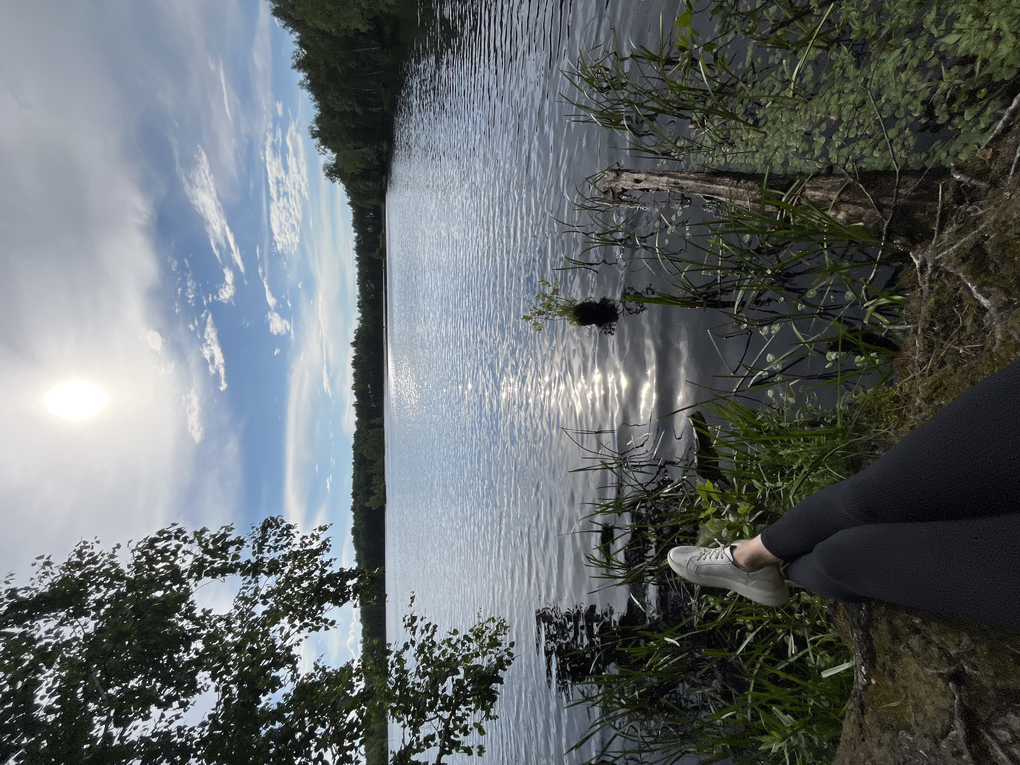 A person's legs, clad in black leggings and white sneakers, are prominently featured in the foreground, resting on a moss-covered surface next to the dark water of a lake. Reeds and other aquatic plants grow along the water's edge, with a weathered wooden log partially submerged. The lake extends into the background, reflecting the bright sun and the sky, which is a mix of blue and white clouds. A line of dense green trees borders the far side of the lake, suggesting a natural, possibly rural, setting. The scene captures a moment of relaxation by the water under a bright, sunny sky. The specific location of Нагорный, Russia, is not discernible from the visual information present in the image.