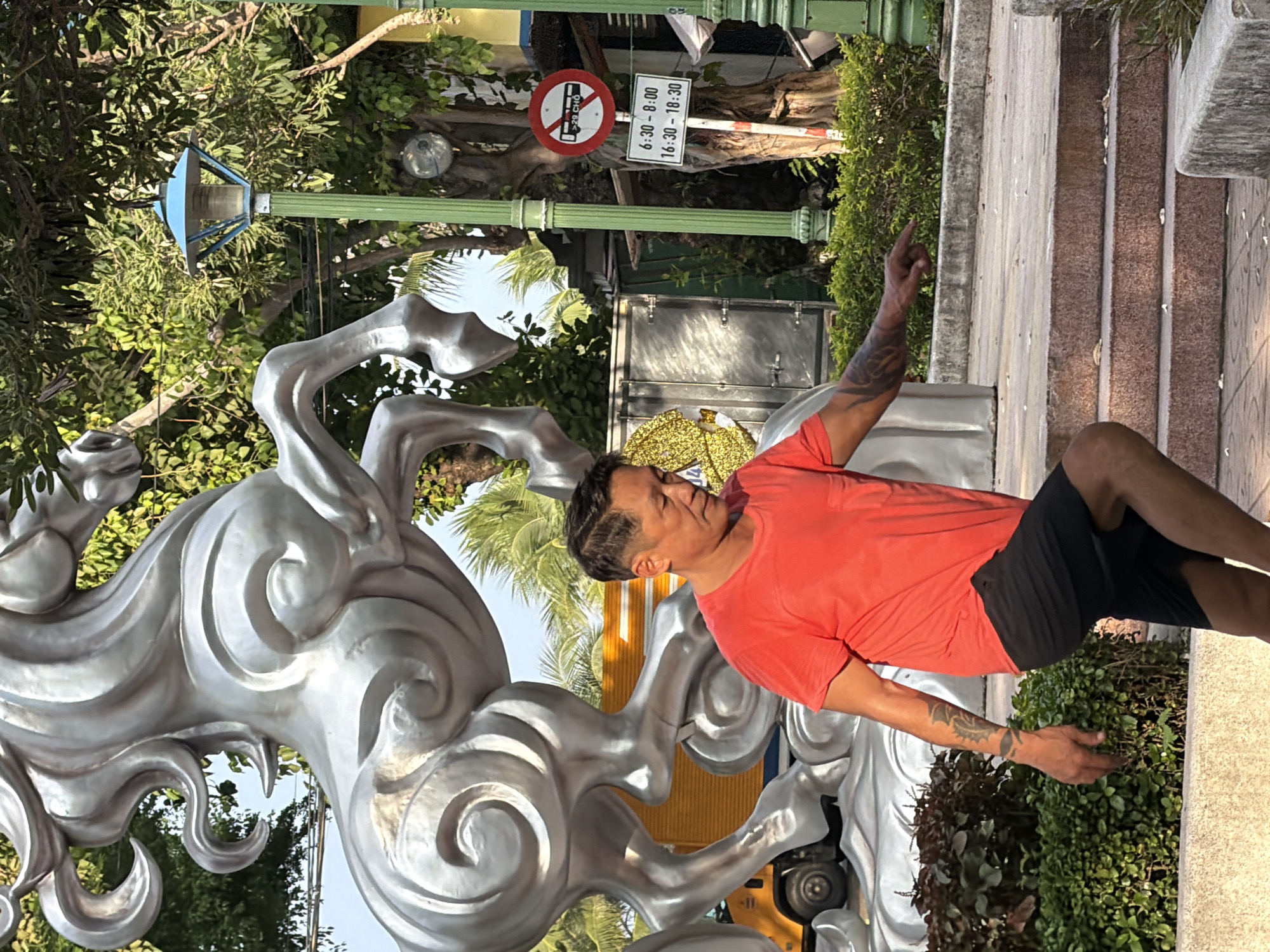 A middle-aged East Asian man with short dark hair, wearing an orange t-shirt and black shorts, walks down a set of stone stairs. He has a dragon tattoo on his left arm and is pointing with his right hand. Behind him, a large, silver, abstract sculpture of a horse is visible, with a yellow truck and a white truck parked behind it. The scene is outdoors, with lush green trees and palm trees in the background. A green lamppost with a blue light fixture stands to the right, next to a sign with a circular red and white symbol indicating no buses and the text "6:30-8:00" and "16:30-18:30". This location is identified as being in the city of Nha Trang, Vietnam.