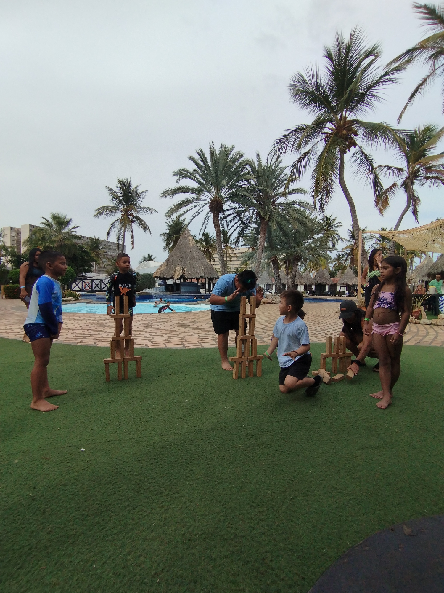In Porlamar, Venezuela, a group of children and adults are playing a game with wooden blocks near a pool and palm trees. One boy is standing on a completed block tower, while others are constructing their own towers. The scene is set on a grassy area, with the pool and tropical vegetation in the background. Several buildings and thatched roof structures are visible in the distance. The sky is overcast.