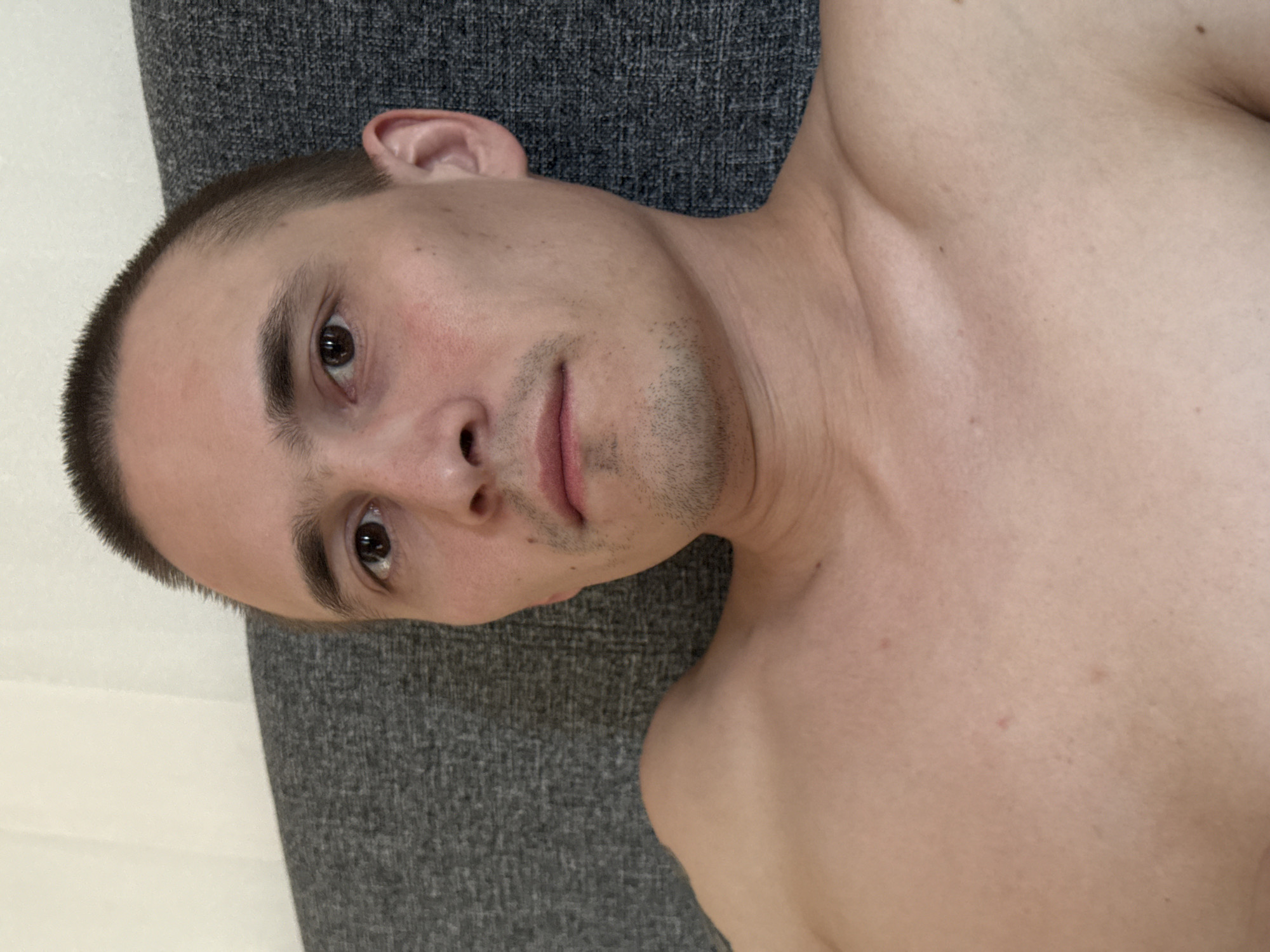 A shirtless man with a shaved head lies on a gray fabric surface. His head is tilted to the left, with his face looking directly at the viewer. He has fair skin, brown eyes, and short stubble on his chin. His right ear is visible. The background includes a neutral-colored wall. The location in Chaikovsky, Russia is not determinable from the image.